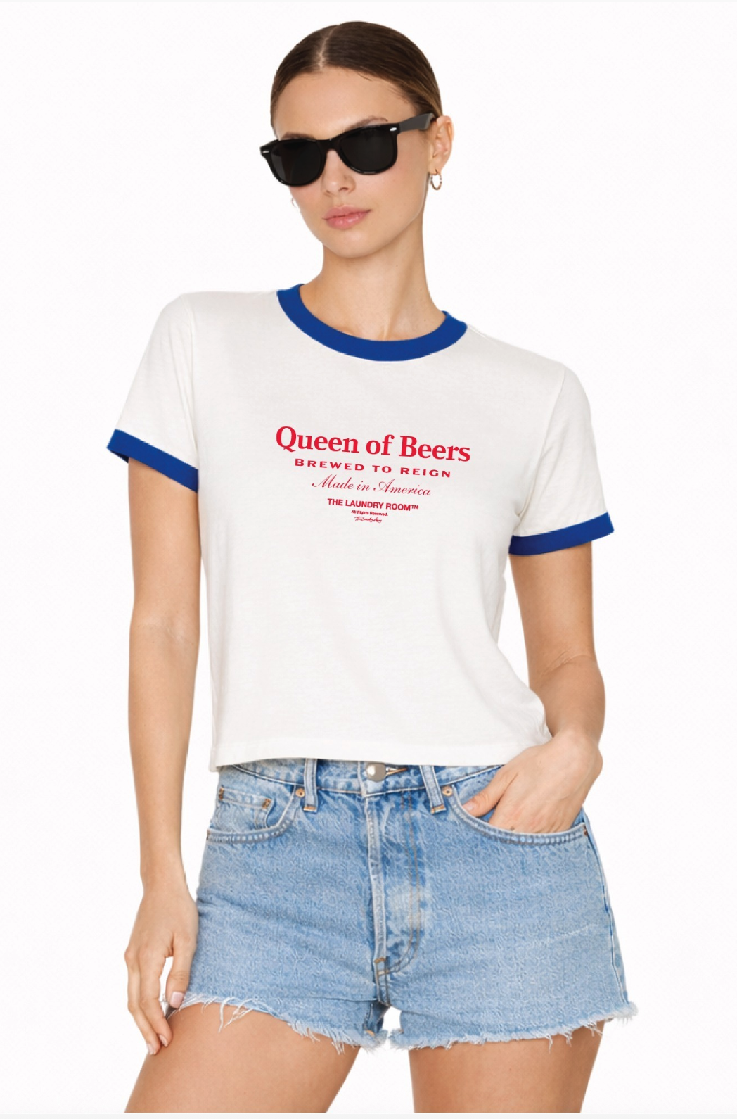 A woman in The Laundry Room Queen Of Beers Ringer Tee in White Multi and denim shorts poses with one hand in her pocket against a plain background, wearing black sunglasses.