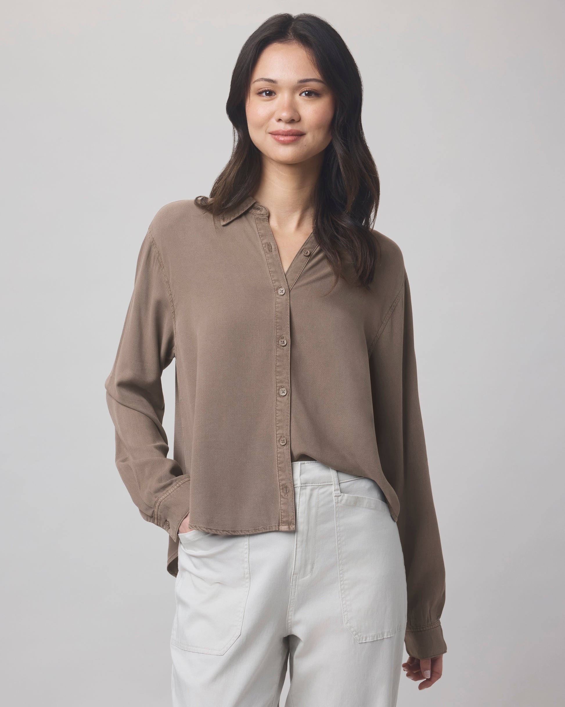 A woman with long dark hair wears the Splendid Joanne Blouse in a relaxed fit with a collar, paired with light gray pants. She stands against a plain light background, smiling softly with one hand in her pocket.