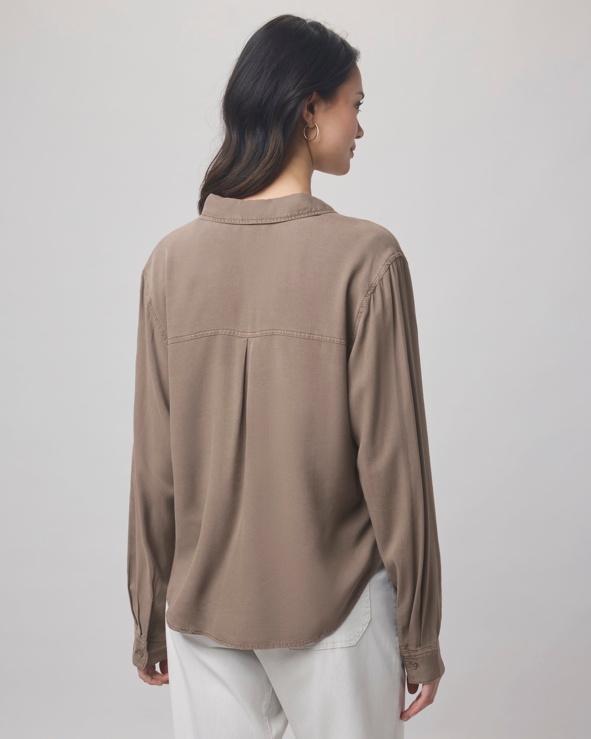 A woman with long dark hair stands with her back to the camera, wearing the Splendid Joanne Blouse in taupe and white pants against a plain, light background.