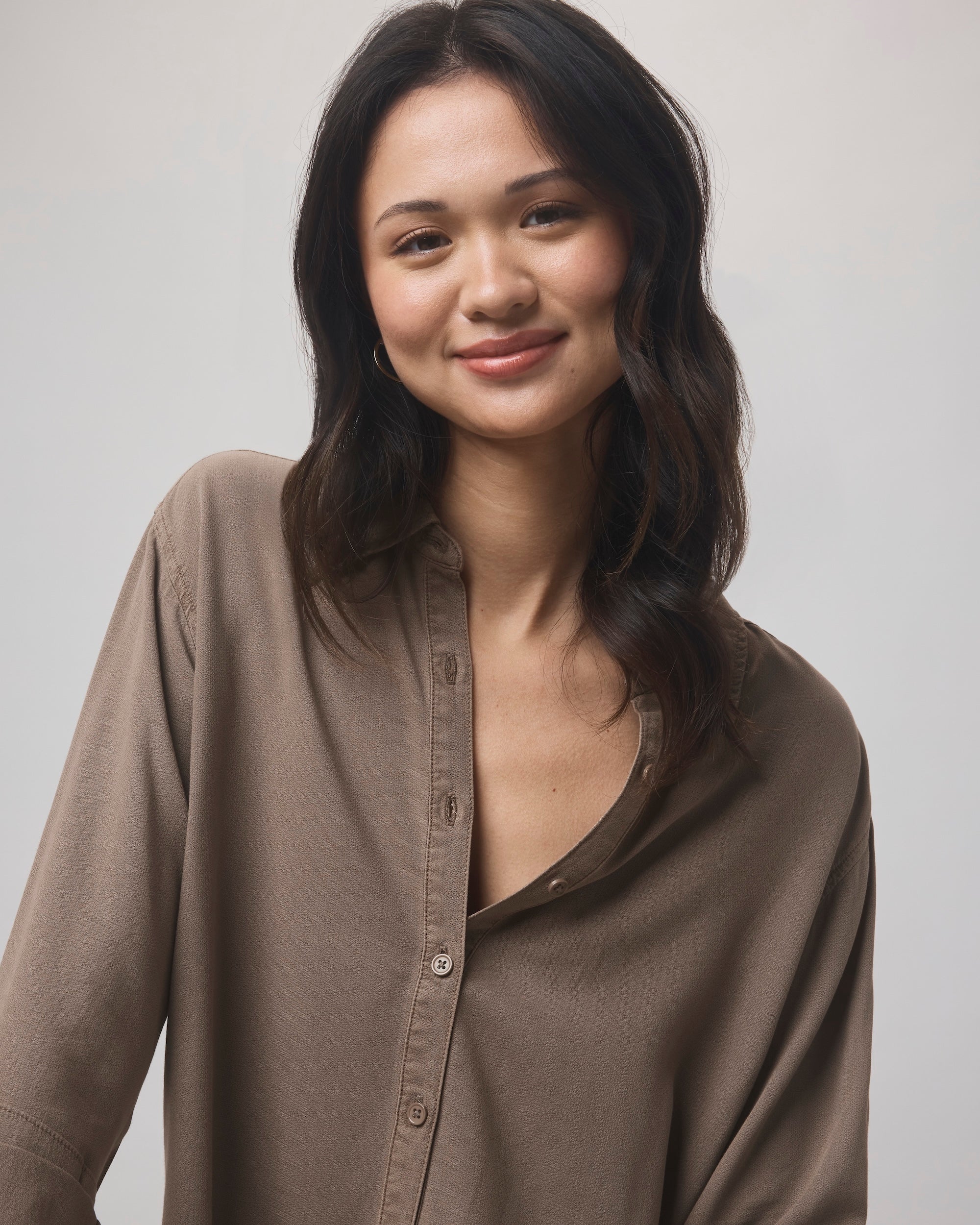 A woman with long, wavy dark hair smiles softly at the camera, wearing the Splendid Joanne Blouse— a relaxed fit, collared style with top buttons undone— set against a light, neutral background.