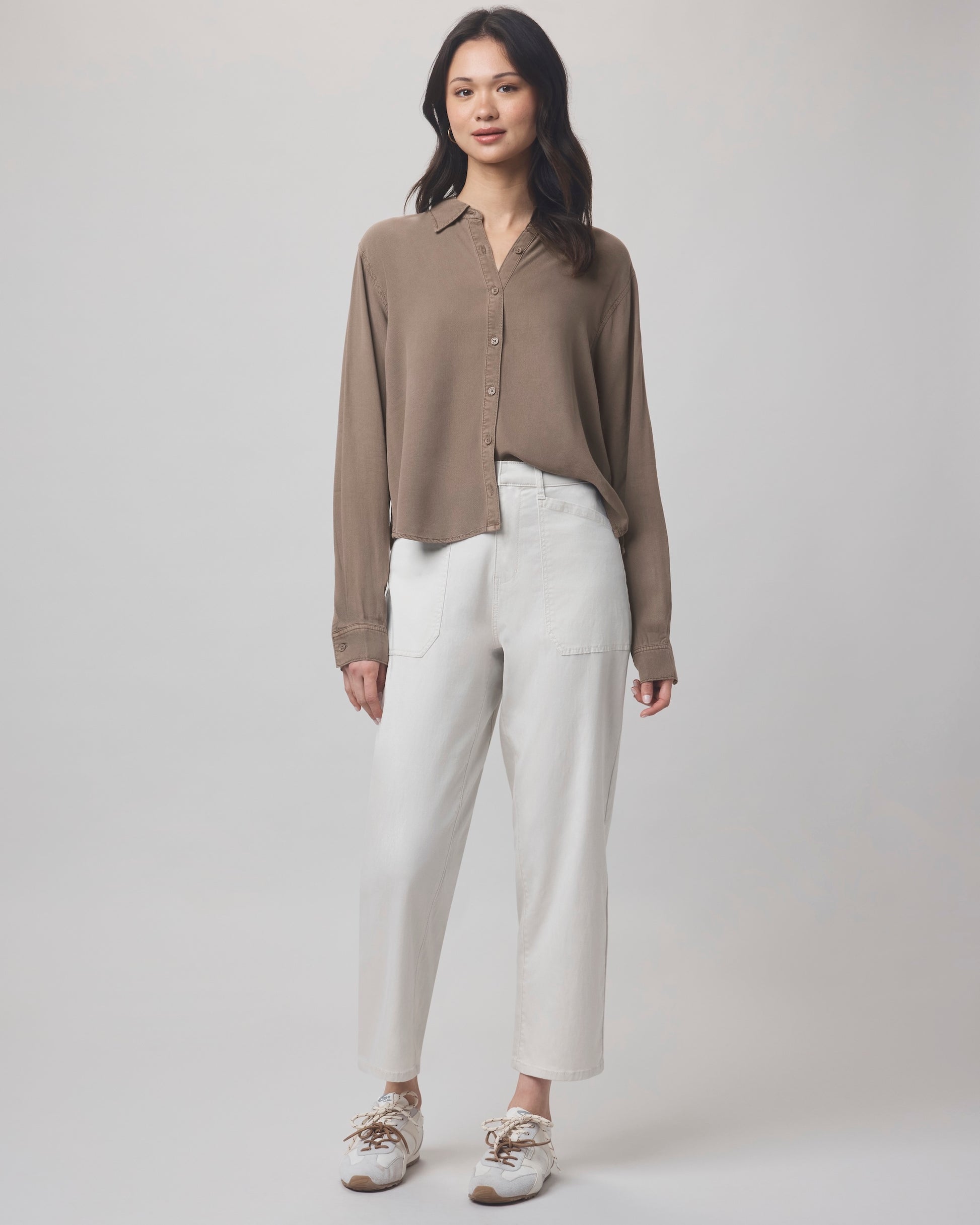A woman with long dark hair and a neutral expression faces forward, wearing the Splendid Joanne Blouse in brown, paired with white high-waisted pants featuring large front pockets and white sneakers, against a light background.