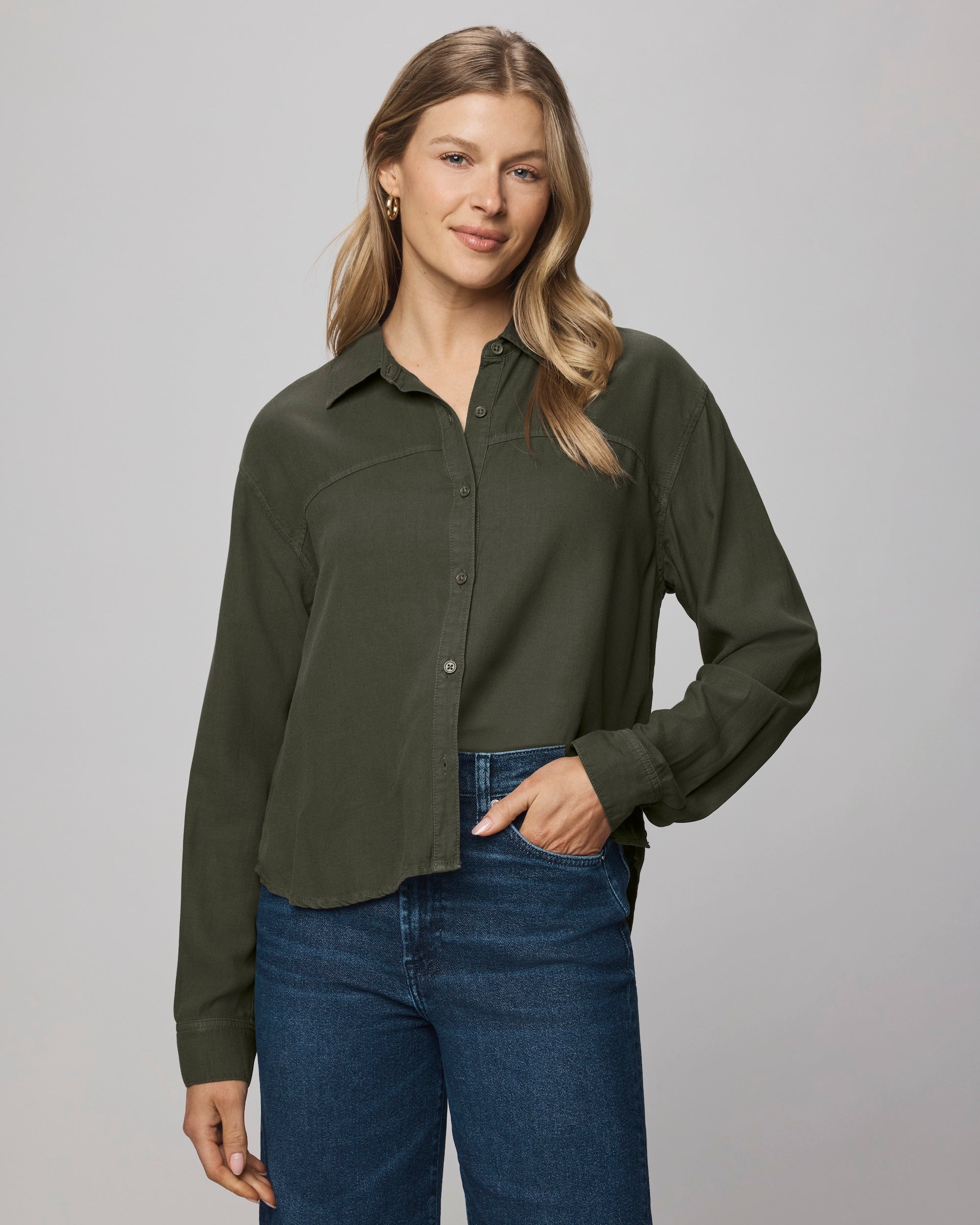 A woman with long blonde hair wears the Splendid Joanne Blouse in dark green and blue jeans, standing against a light gray background and smiling with one hand in her pocket.