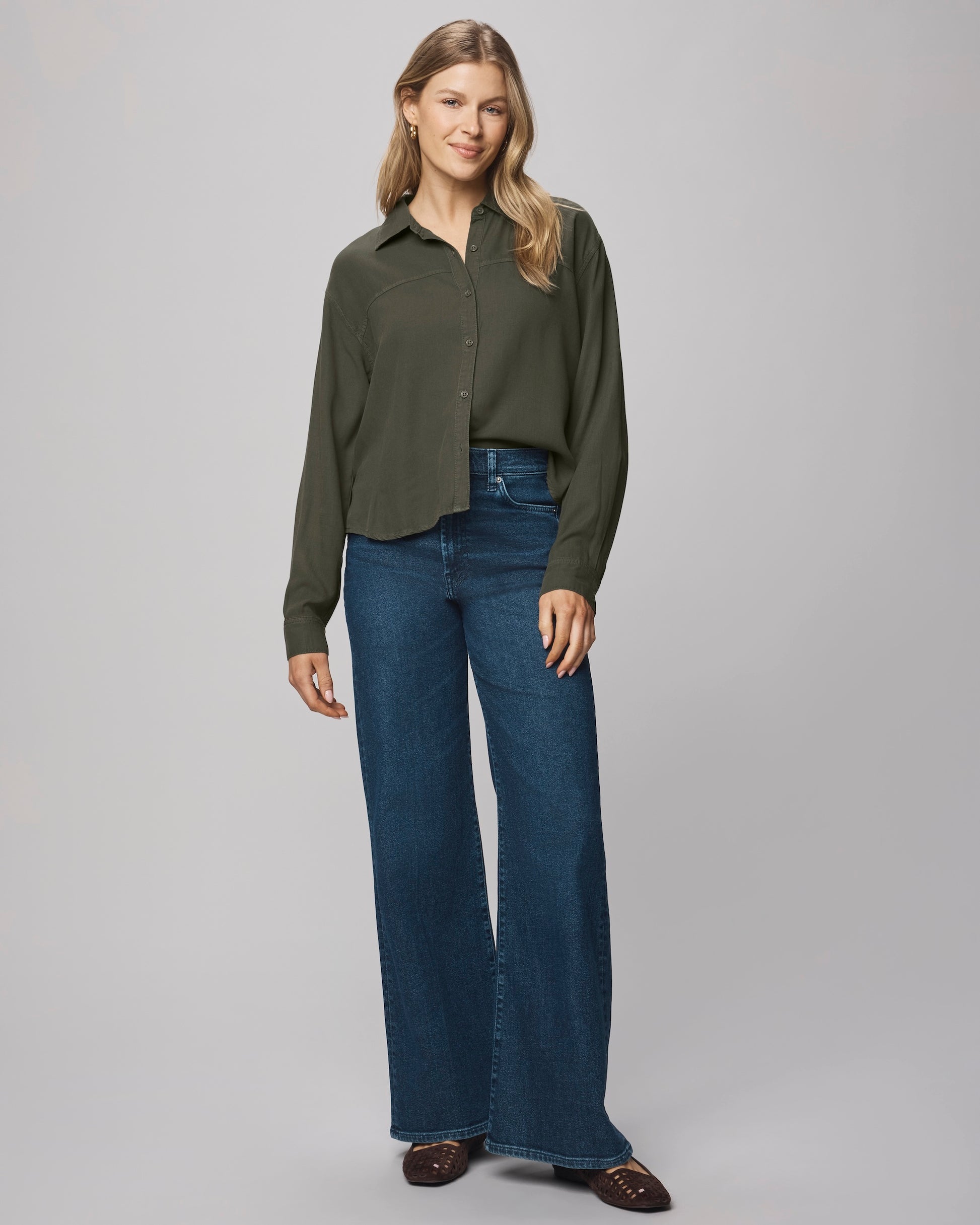 A woman with light brown, wavy hair smiles softly as she models the Splendid Joanne Blouse—a dark green, relaxed fit collared top—paired with high-waisted, wide-leg blue jeans and brown shoes against a plain background.