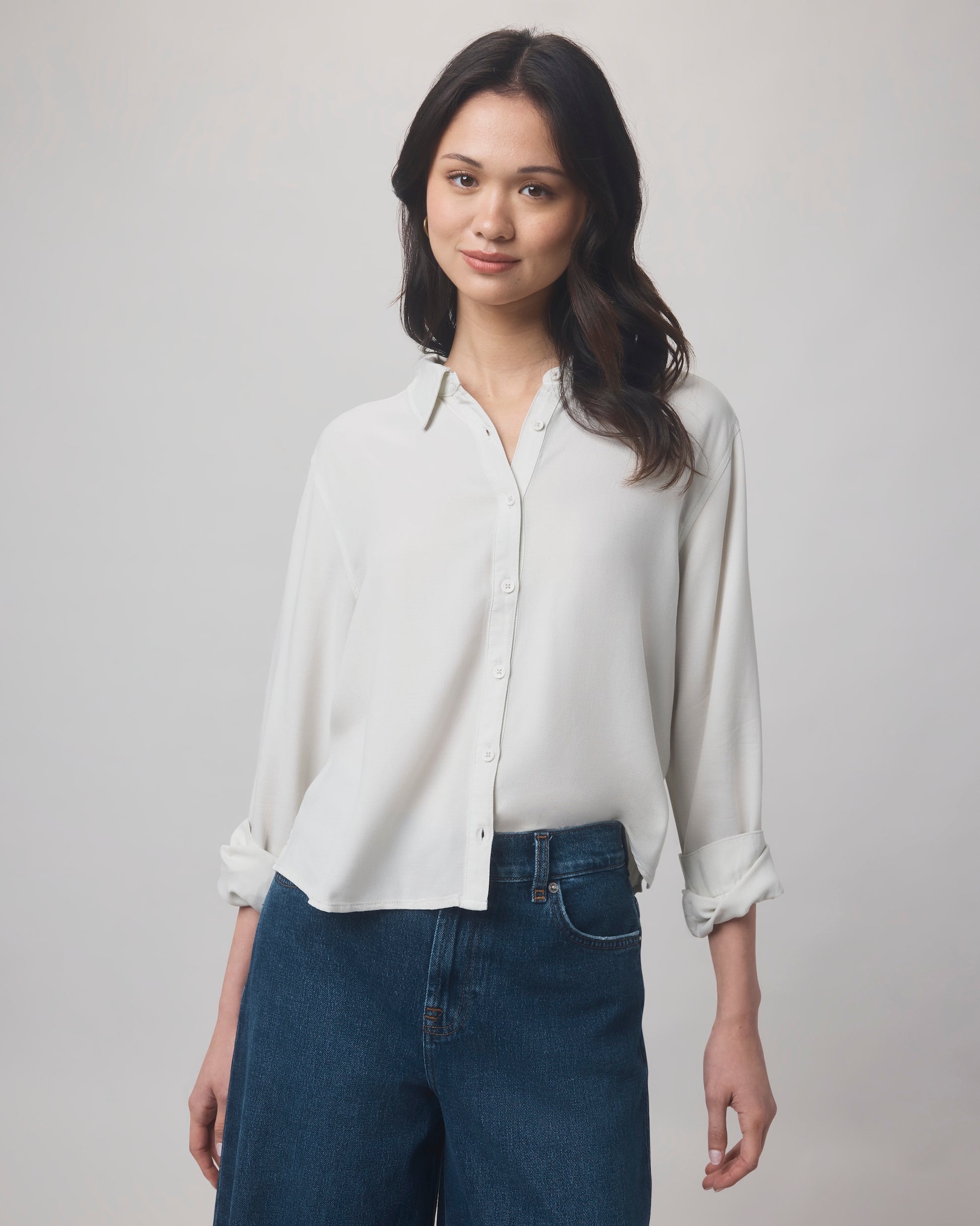 A woman with long dark hair wears the Splendid Joanne Blouse, a white relaxed-fit top with rolled sleeves, paired with blue jeans. She stands against a plain light background and smiles slightly at the camera.