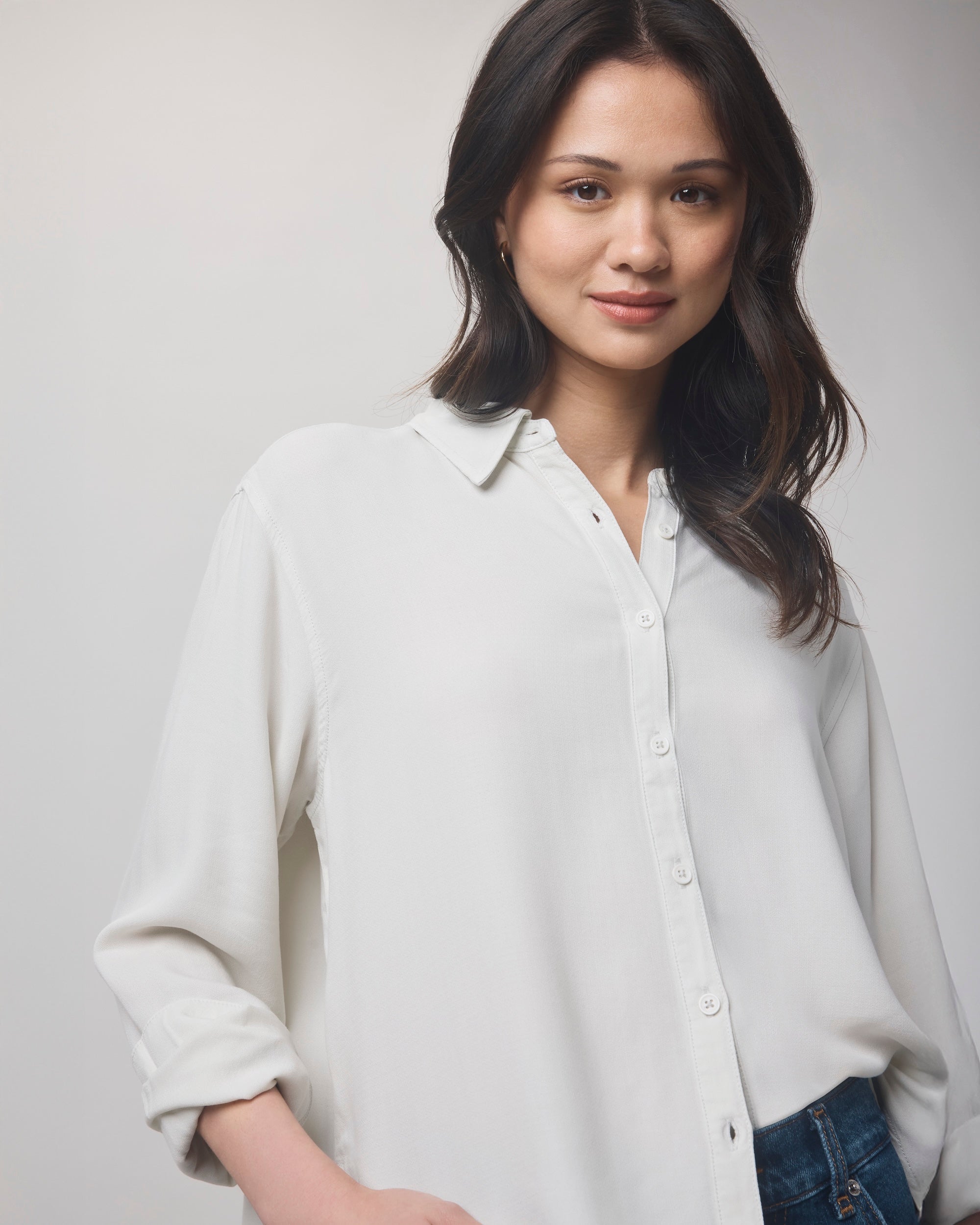 A woman with long, dark hair models the Splendid Joanne Blouse, a relaxed-fit top, standing against a light gray background and smiling gently at the camera with one hand in her pocket.