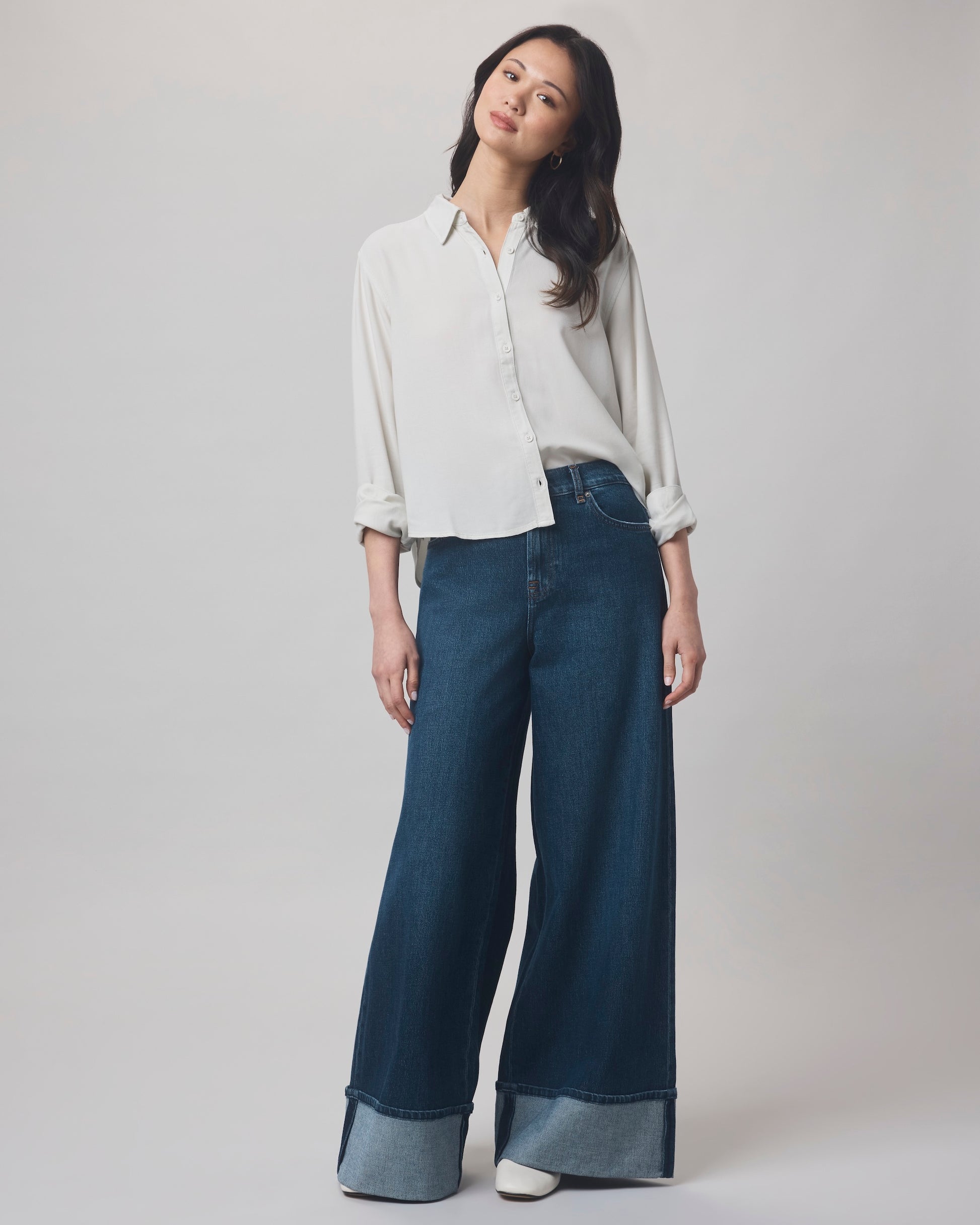 A woman poses against a plain background in the Splendid Joanne Blouse, a relaxed-fit white button-up with rolled sleeves, paired with high-waisted wide-leg blue jeans with large cuffs. She smiles softly with her head slightly tilted.