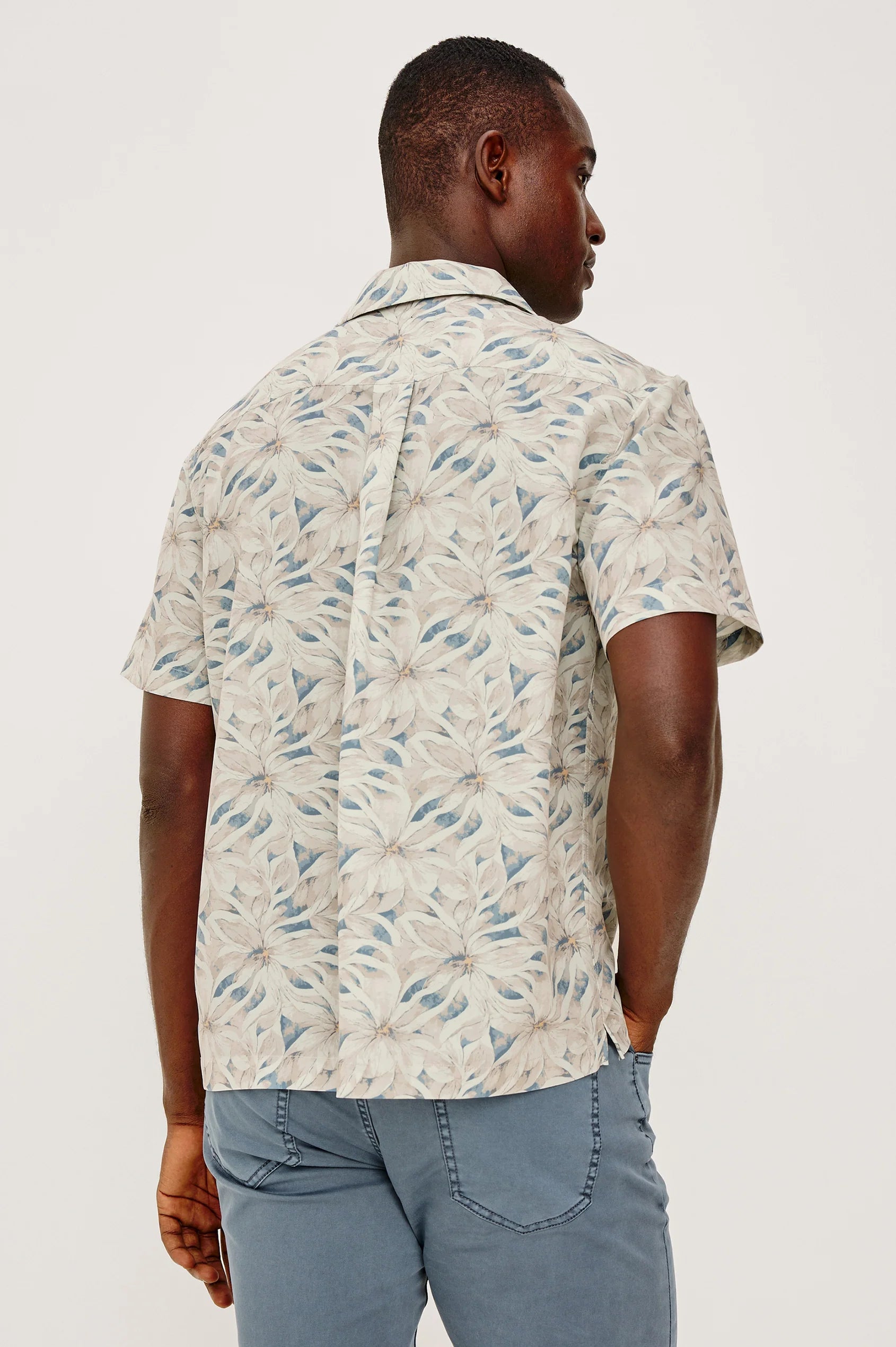 A man faces away wearing the Rails Rhodes Shirt, a vintage-inspired short-sleeve cropped button-up with a beige and blue leaf print, paired with light blue pants. The background is plain and light-colored.