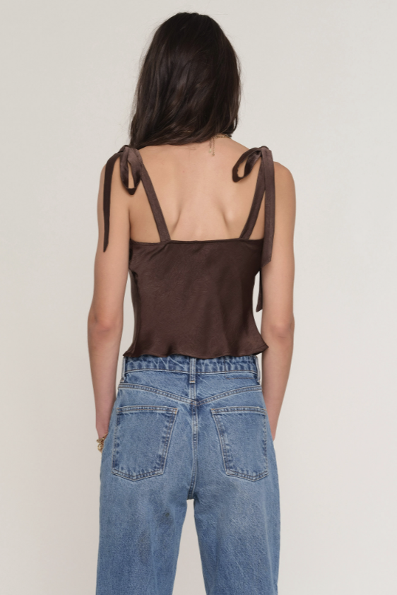 A woman with long brown hair is shown from the back, wearing the Heartloom Ricki Top—a brown satin sleeveless top with adjustable straps—and high-waisted blue jeans, set against a plain light background.
