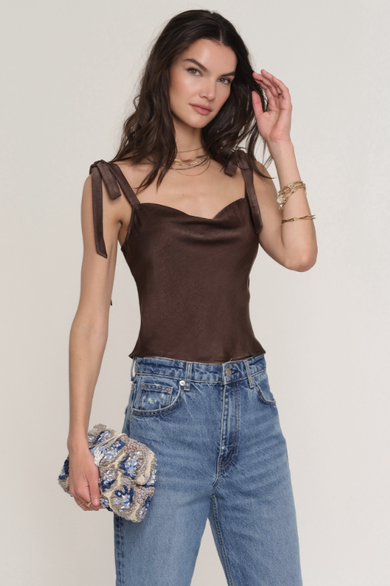 A woman with long, wavy dark hair wears the Heartloom Ricki Top in brown—a satin tank with adjustable straps and a cowl neckline—paired with blue jeans, layered necklaces, bracelets, and a textured clutch bag.