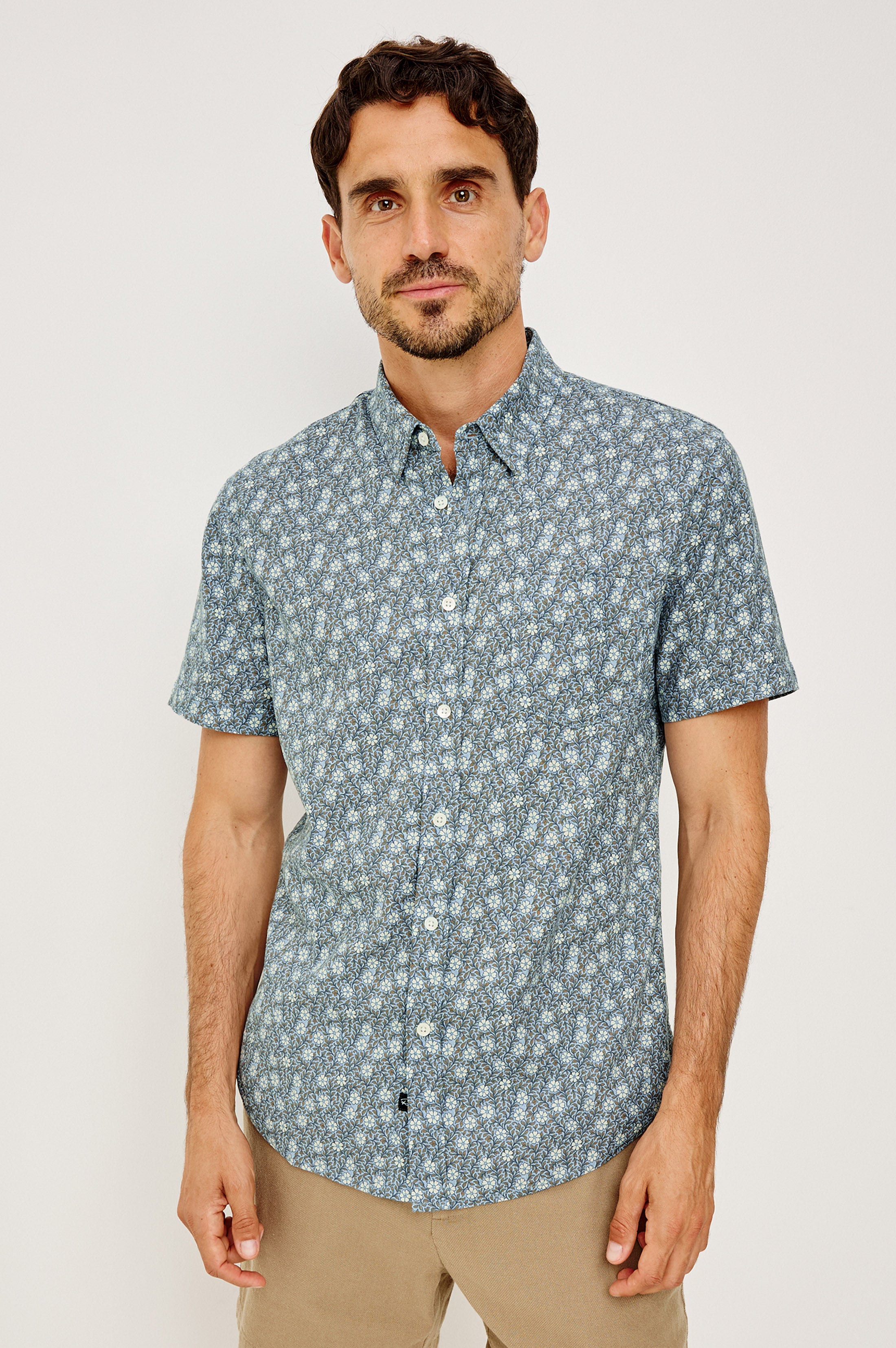 A man with short dark hair and a trimmed beard wears the Rails Carson shirt in Blue Multi, featuring short sleeves and a patterned denim look, paired with beige pants against a plain light background.