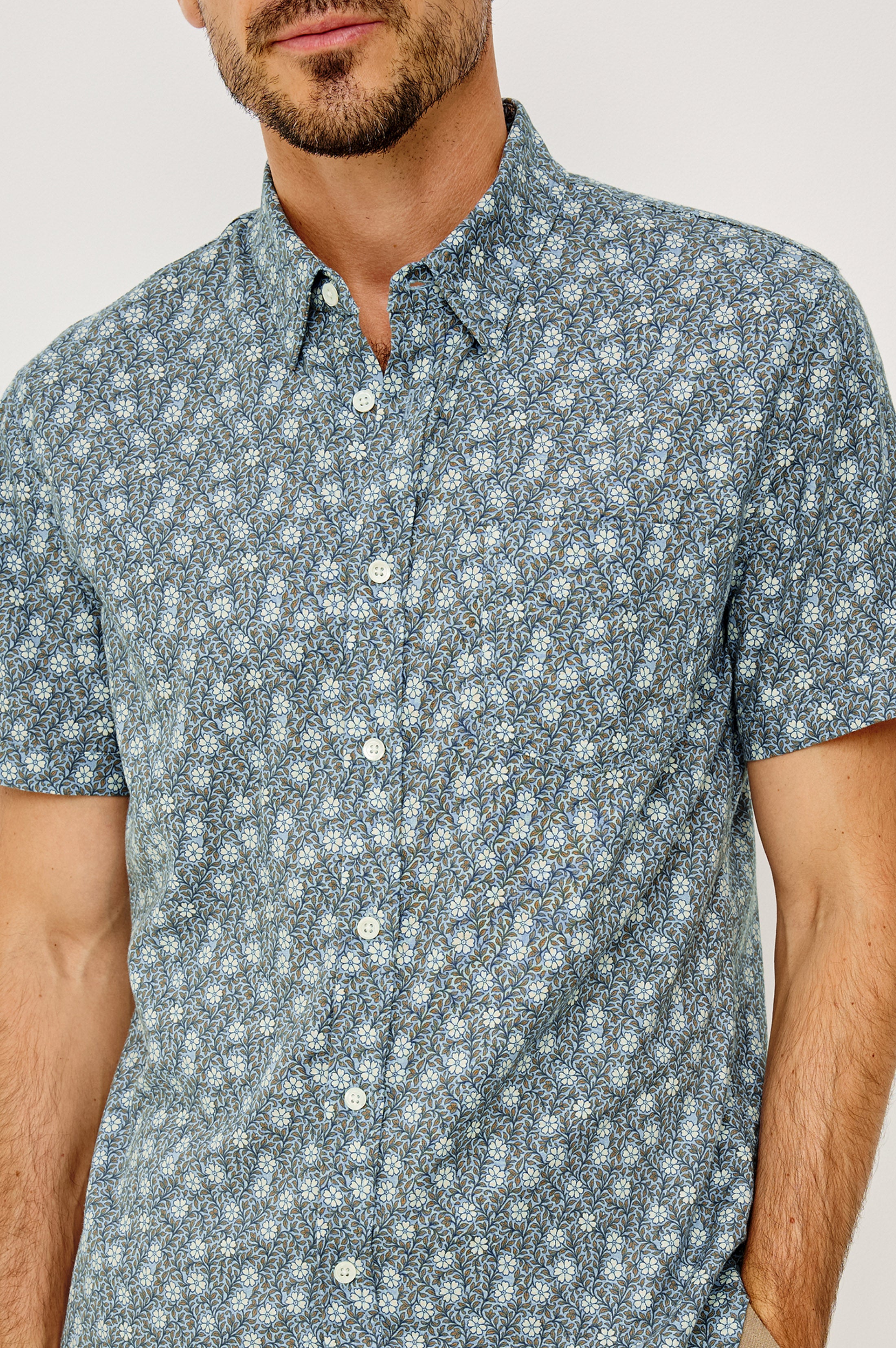 A man wears the Carson short-sleeve blue and white floral linen shirt from Rails, standing against a plain light background with his face partially visible.