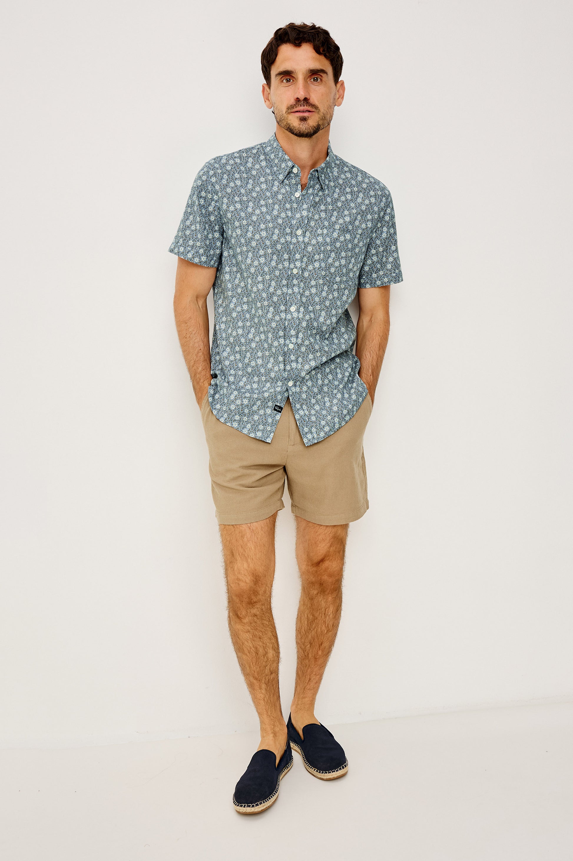 A man with short dark hair and a beard stands against a white wall wearing the Rails Carson short-sleeve linen shirt with a blue pattern, tan shorts, and navy slip-on shoes, his hands in his pockets.