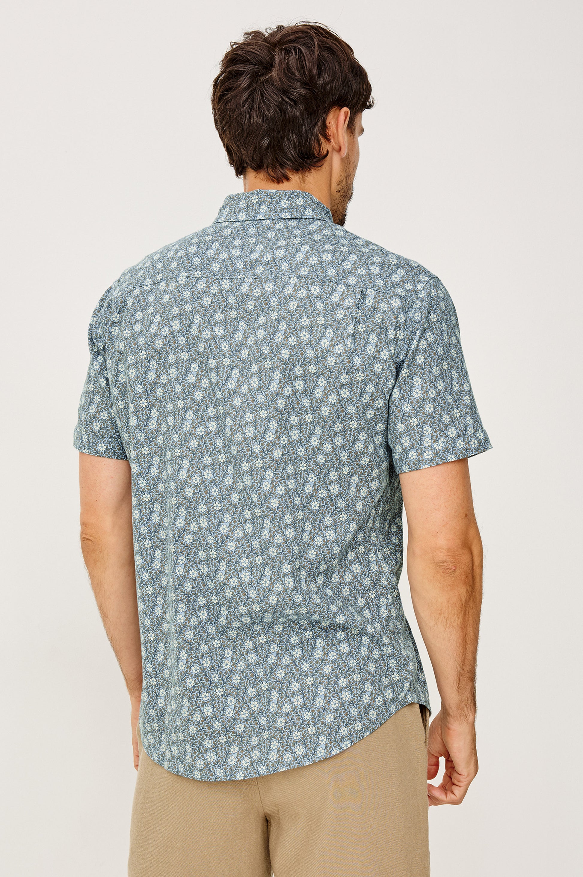 A man faces away, wearing the Rails Carson shirt—a light blue short sleeve top with a small floral pattern—and beige pants. The background is plain and light-colored.