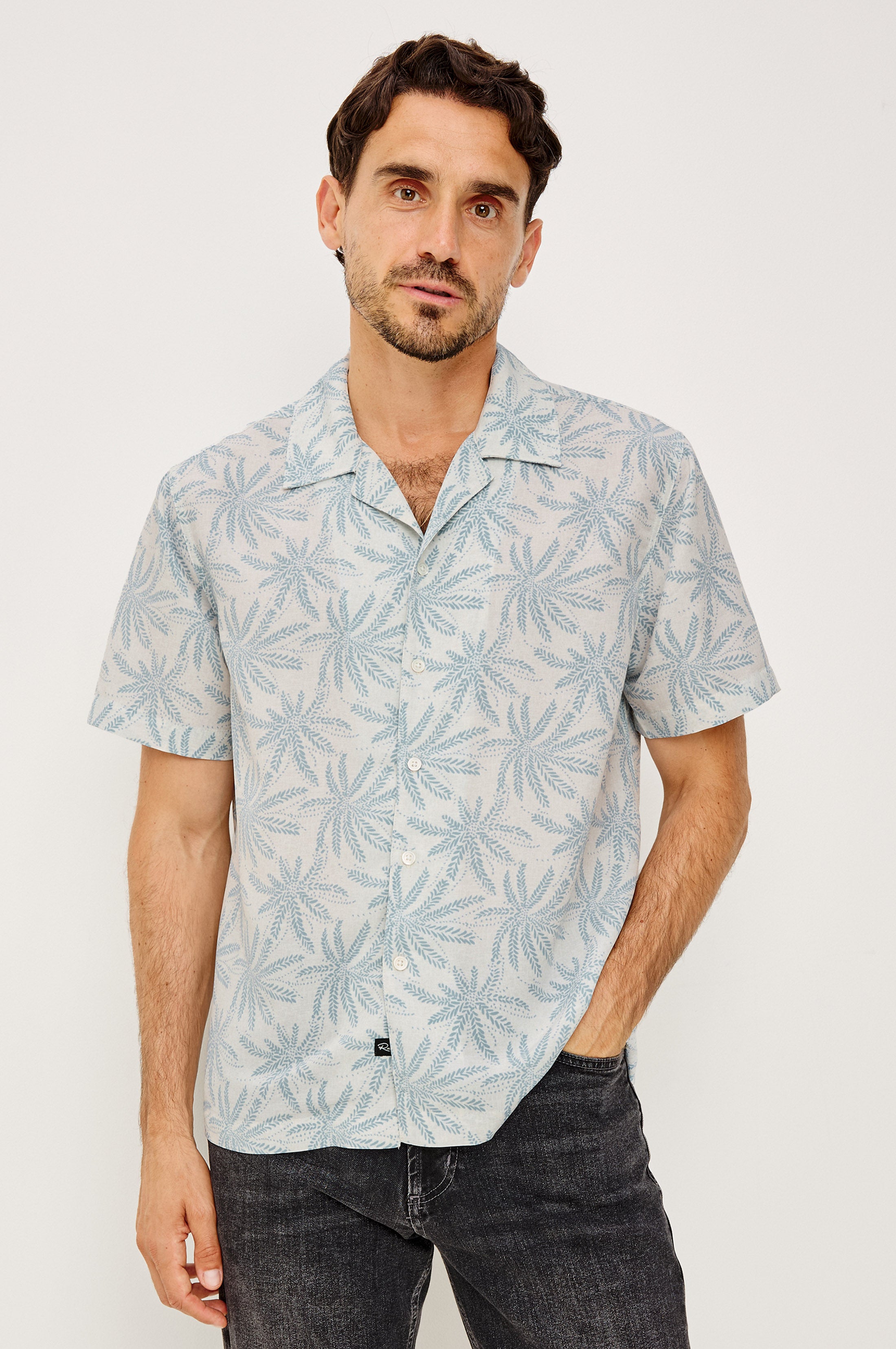 A man with short brown hair and a trimmed beard wears the Rails Rhodes shirt in Blue Multi, featuring a drapey crepe fabric, vintage-inspired notched collar, and palm leaf pattern, paired with dark jeans against a white background.