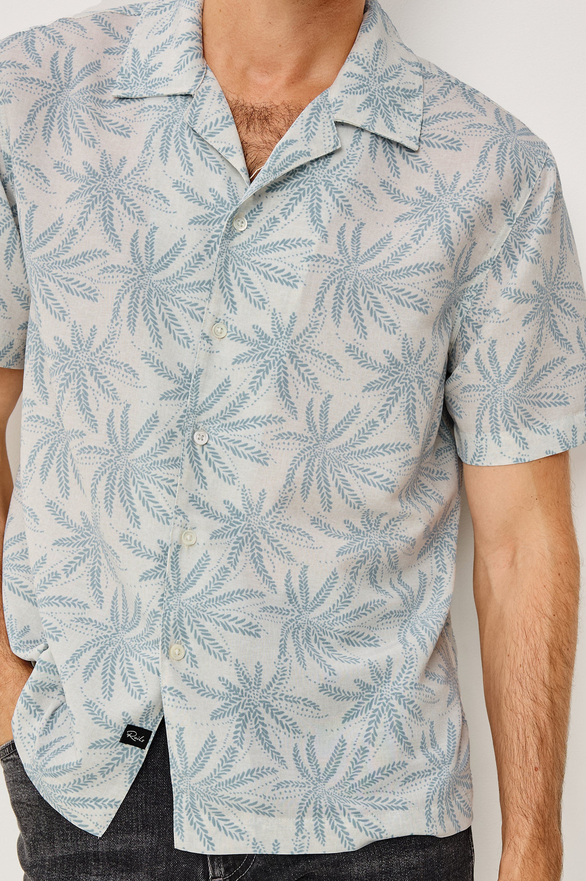 A man wears the Rails Rhodes shirt, a boxy cropped button-up with a blue palm leaf pattern and vintage-inspired notched collar, standing with one hand in his pocket. His head is partially out of frame.