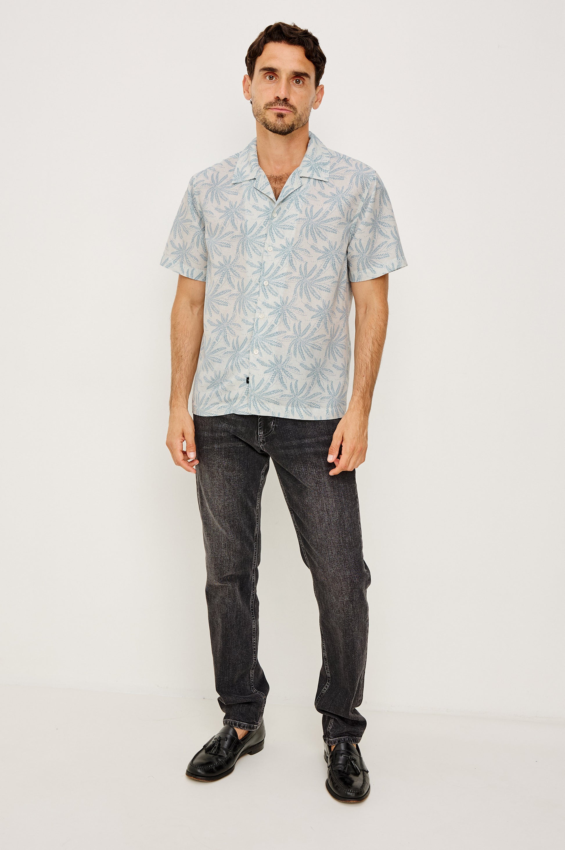 A man with short brown hair and a trimmed beard stands against a white background wearing the Rhodes patterned shirt by Rails, paired with dark jeans and black shoes. He has a neutral expression.