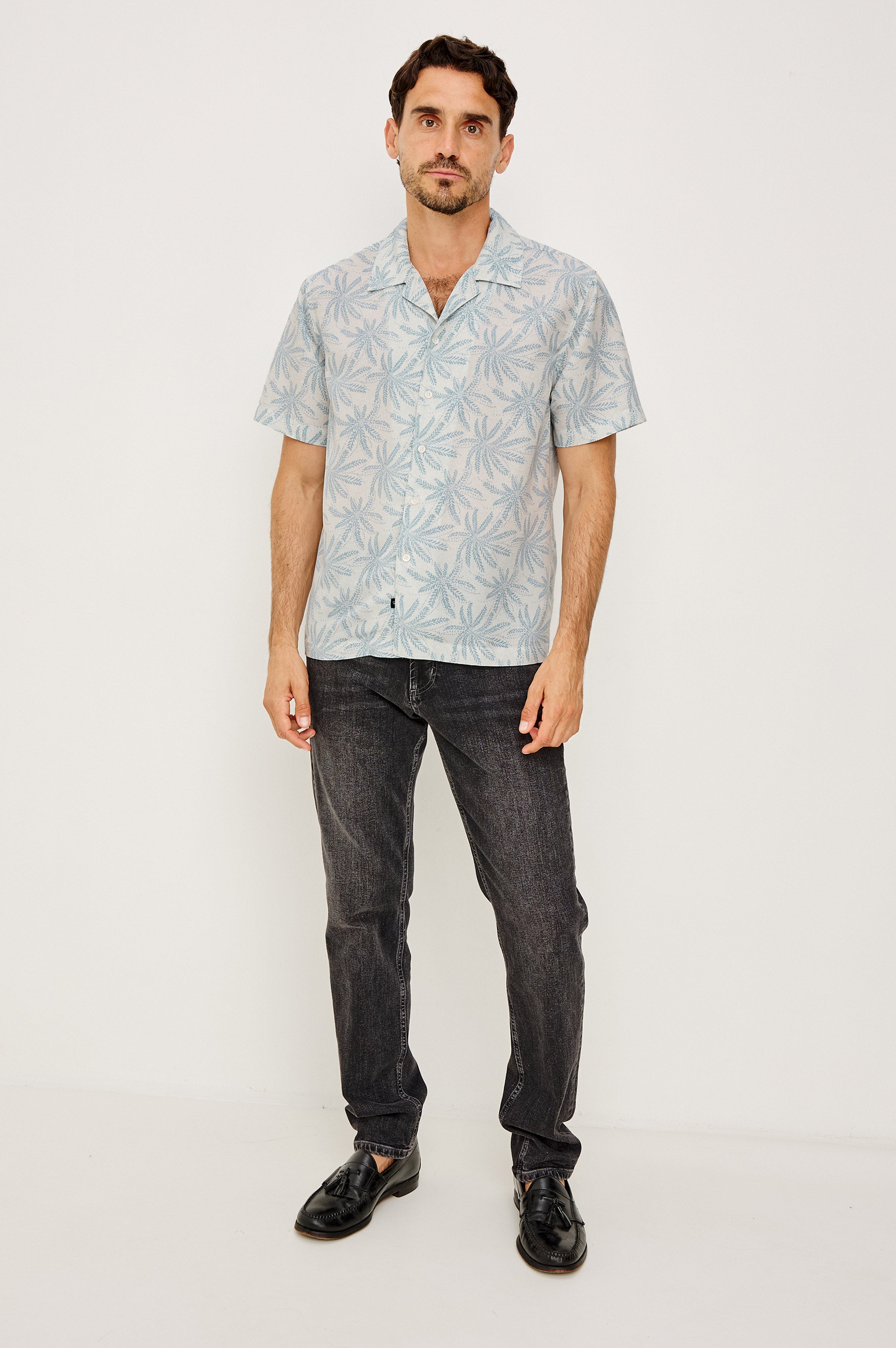 A man with short brown hair and a trimmed beard stands against a white background wearing the Rhodes patterned shirt by Rails, paired with dark jeans and black shoes. He has a neutral expression.