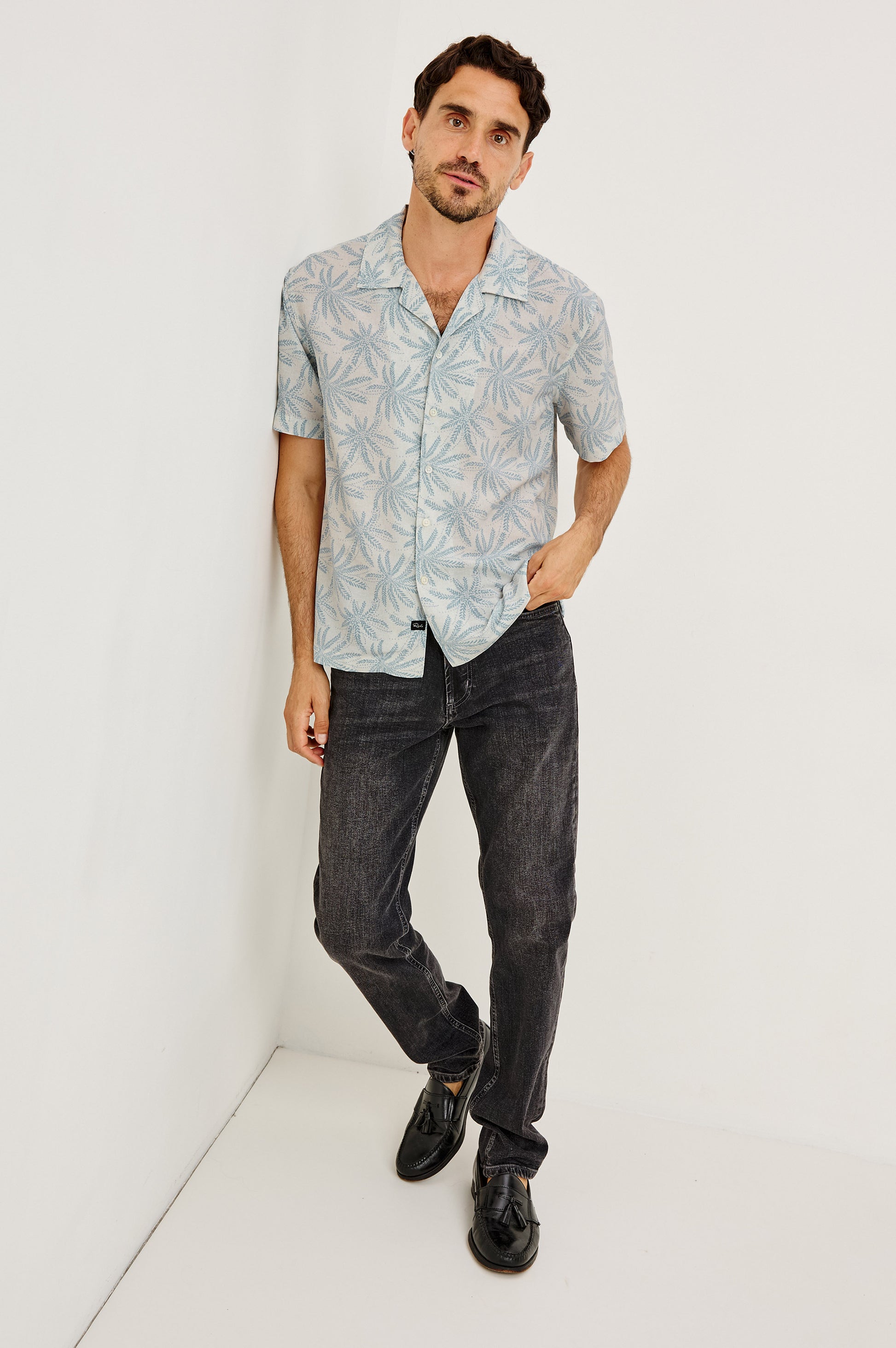 A man with short dark hair and a beard stands against a white wall, wearing the Rhodes light blue patterned boxy cropped button-up shirt by Rails, paired with dark jeans and black shoes. One hand is in his pocket as he looks at the camera.