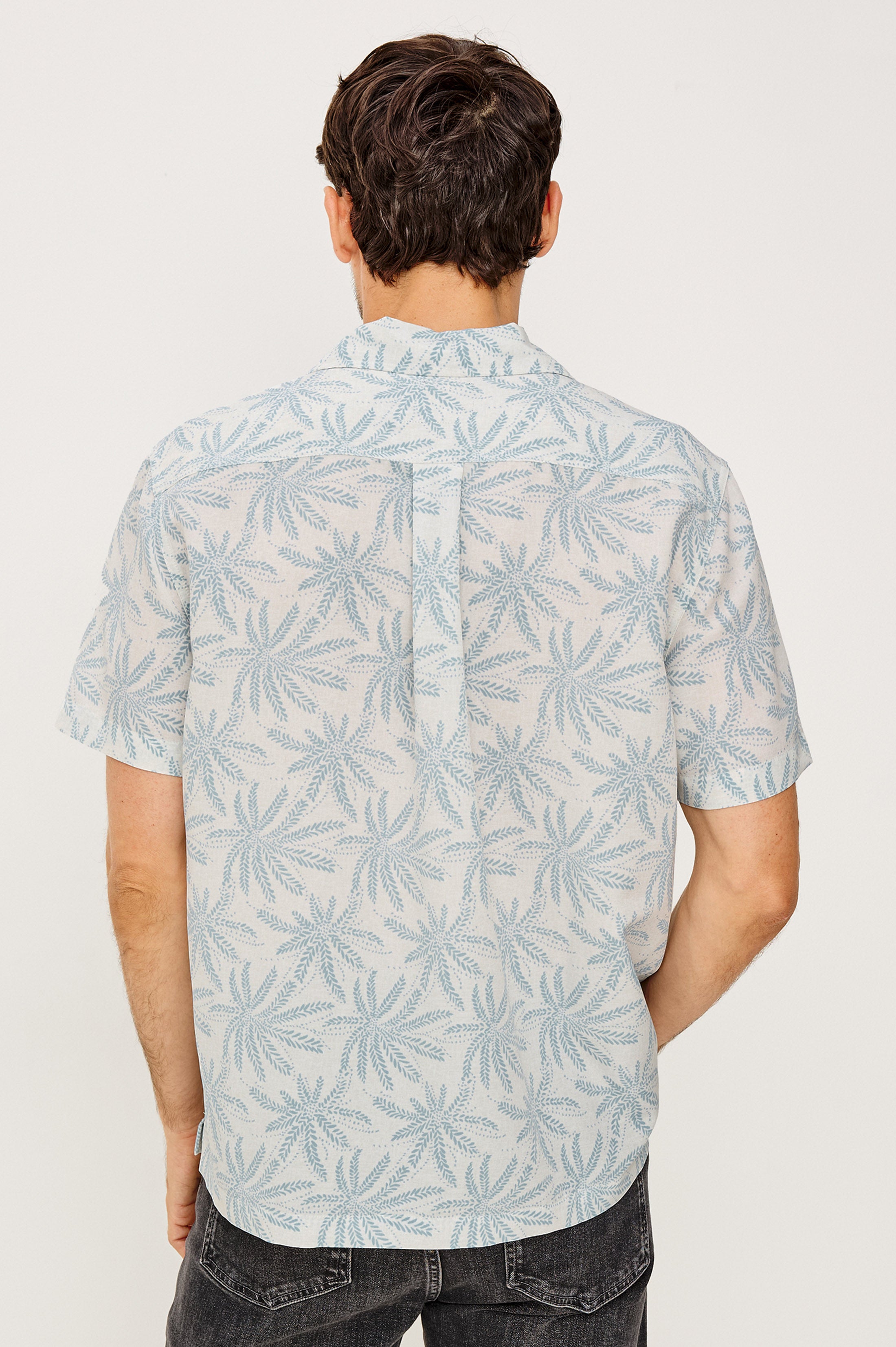 A man with dark hair is seen from the back, wearing the Rails Rhodes vintage-inspired light blue and white leafy notched collar cropped shirt with dark jeans against a plain light background.