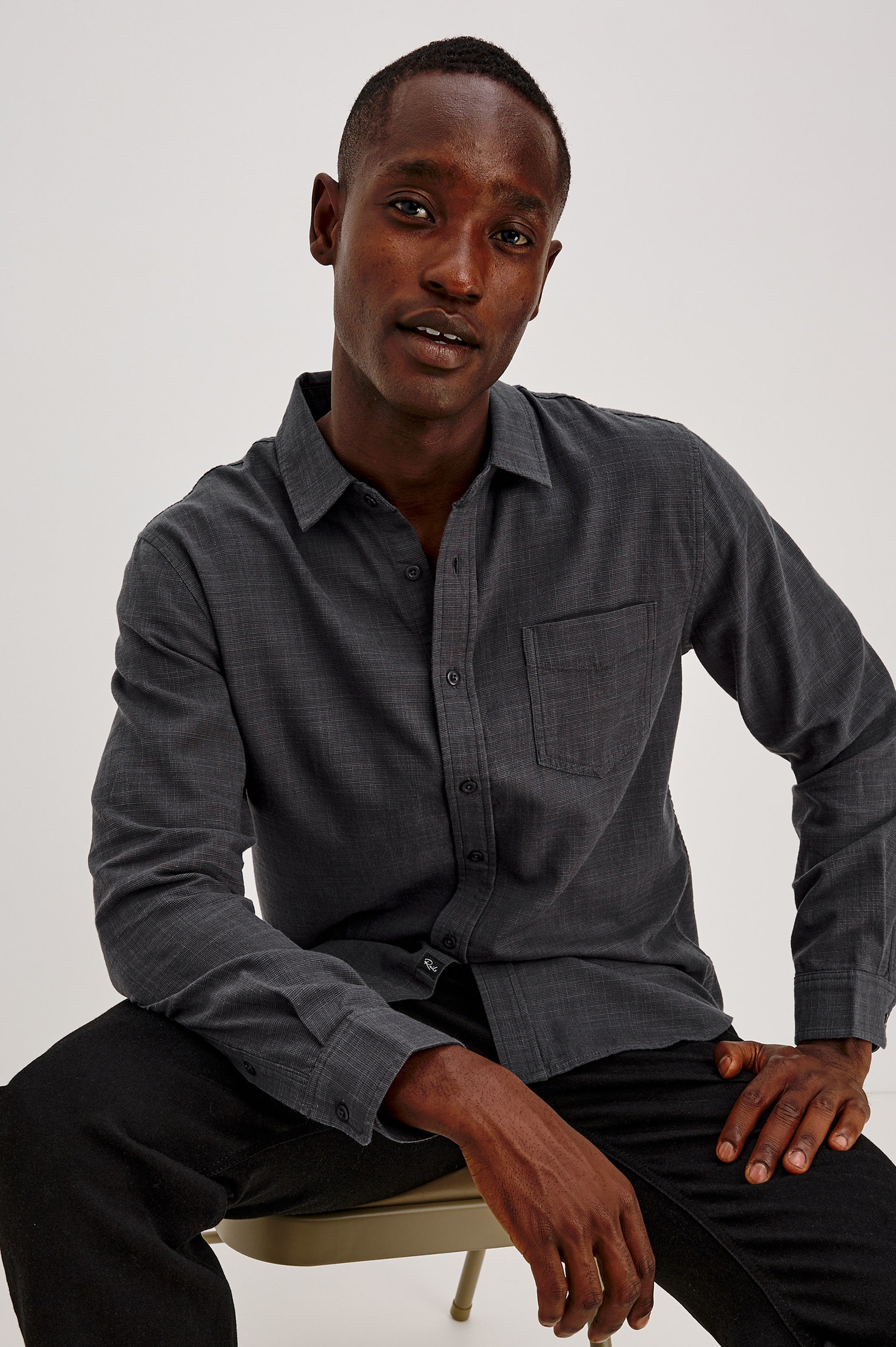 A person wearing the Wyatt Long Sleeve Button Down by Rails in MIDNIGHT ETCH sits on a chair, slightly leaning forward with one hand on their thigh and the other resting on the chair. The background is plain and light-colored.