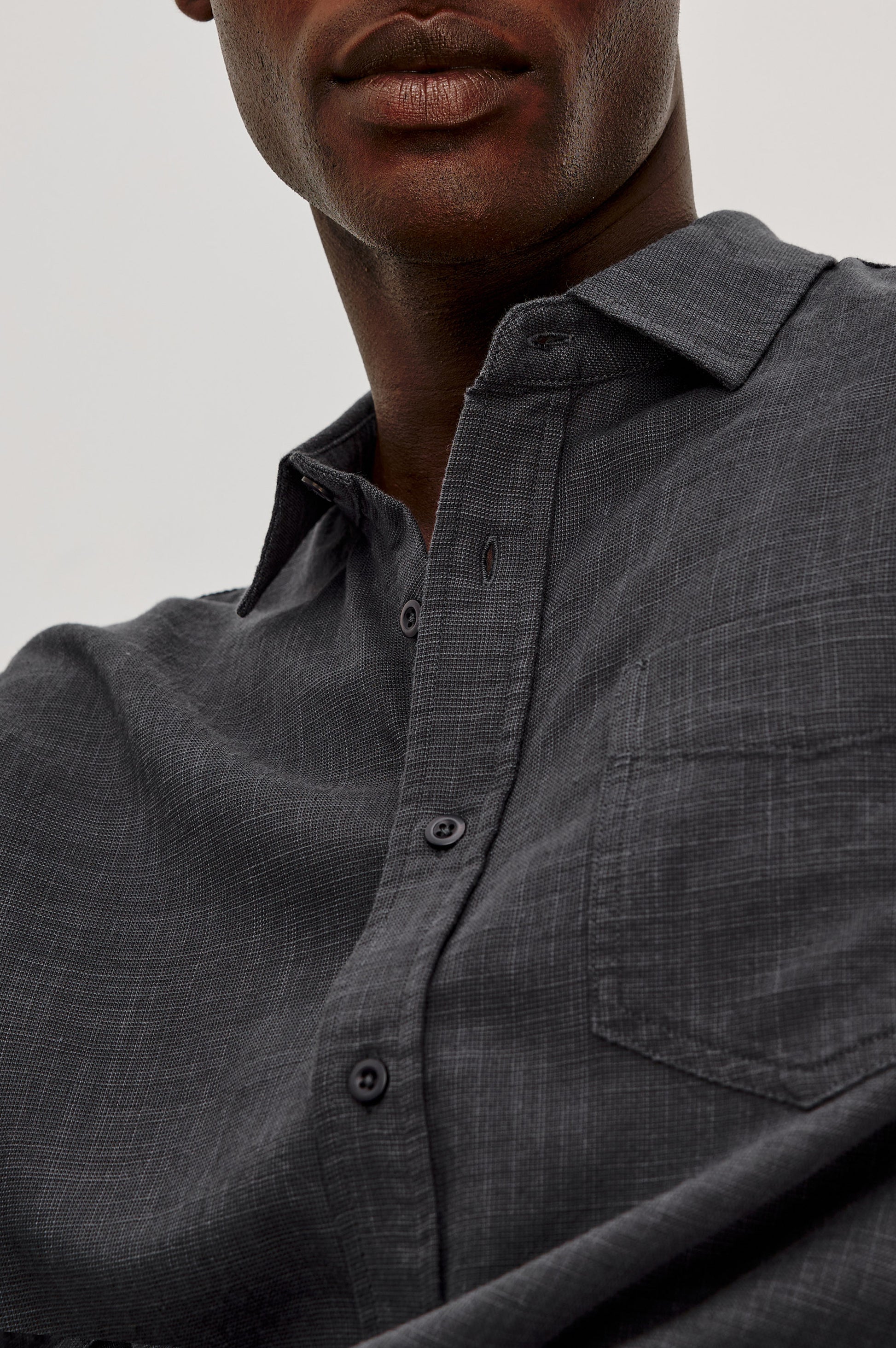 A close-up of a person wearing the Wyatt Long Sleeve Button Down by Rails—a relaxed fit, dark gray cotton shirt featuring a subtle plaid pattern, chest pocket, and button-up front; the person's face is partially visible.