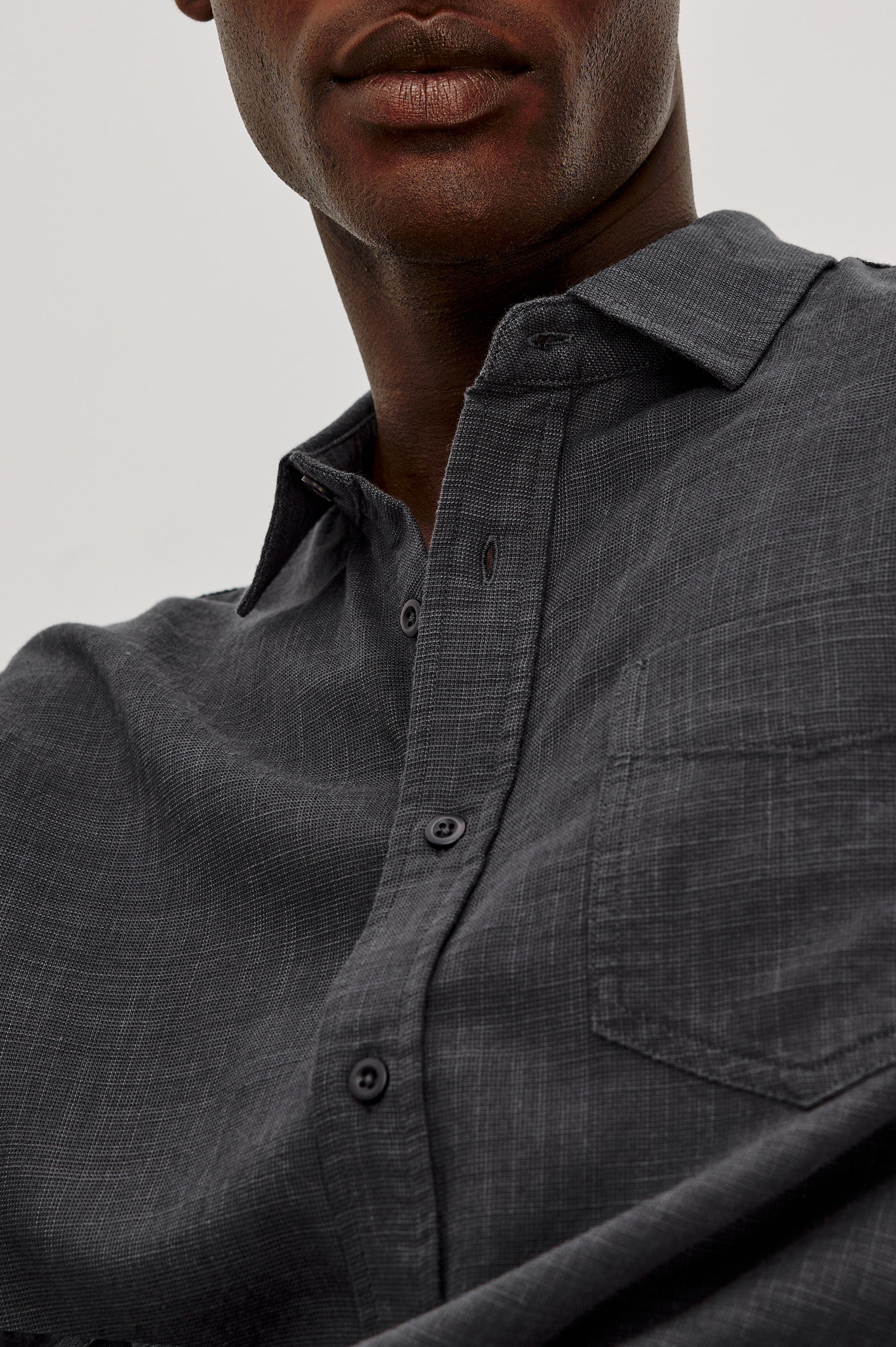 A close-up of a person wearing the Wyatt Long Sleeve Button Down by Rails—a relaxed fit, dark gray cotton shirt featuring a subtle plaid pattern, chest pocket, and button-up front; the person's face is partially visible.