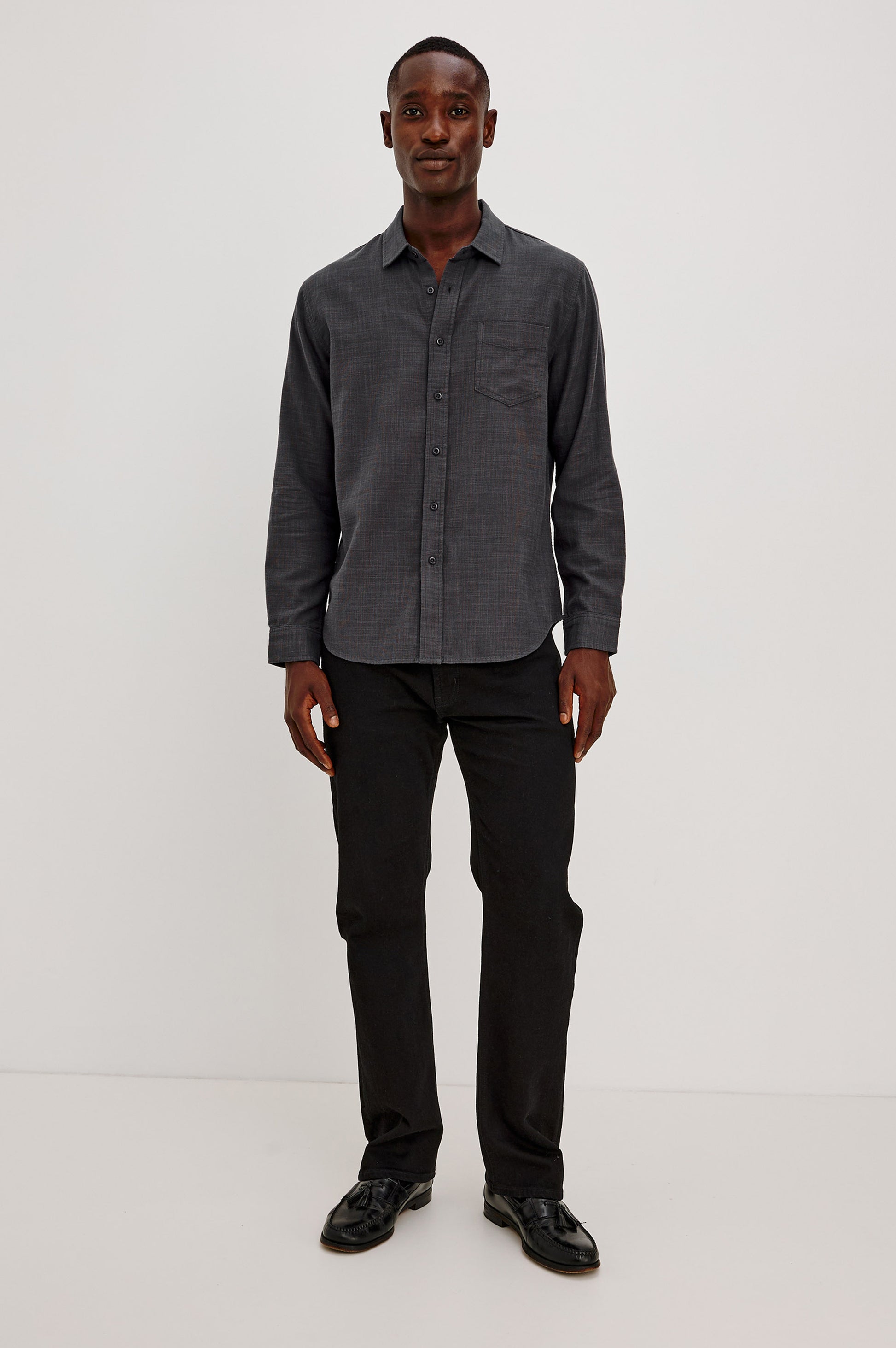 A man stands against a plain white background, wearing the Rails Wyatt Long Sleeve Button Down shirt in dark gray, paired with black pants and black shoes. He is facing forward and smiling.