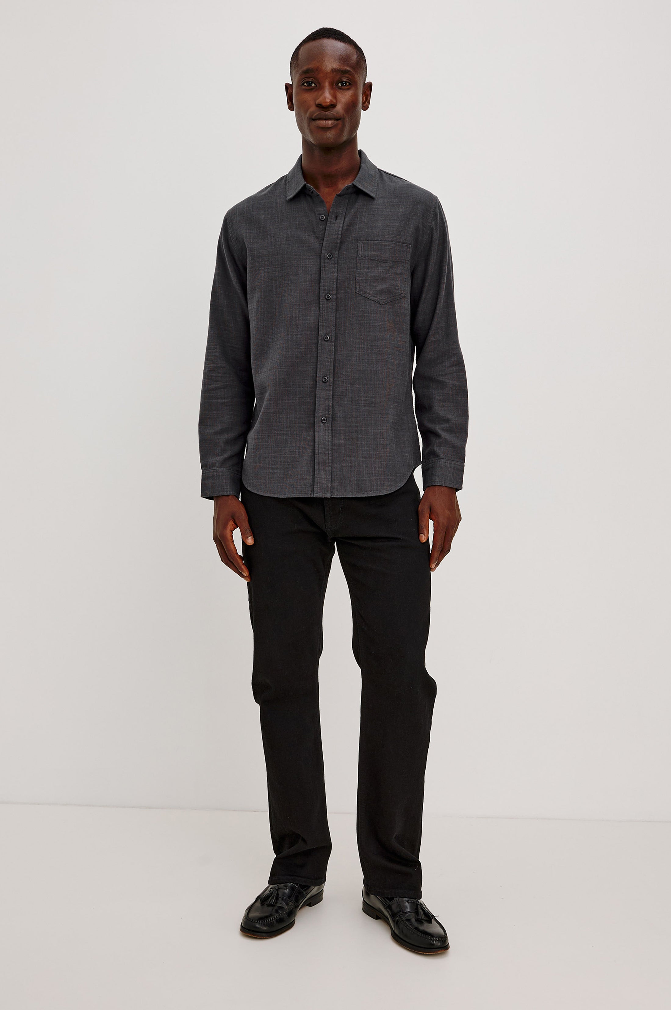 A man stands against a plain white background, wearing the Rails Wyatt Long Sleeve Button Down shirt in dark gray, paired with black pants and black shoes. He is facing forward and smiling.