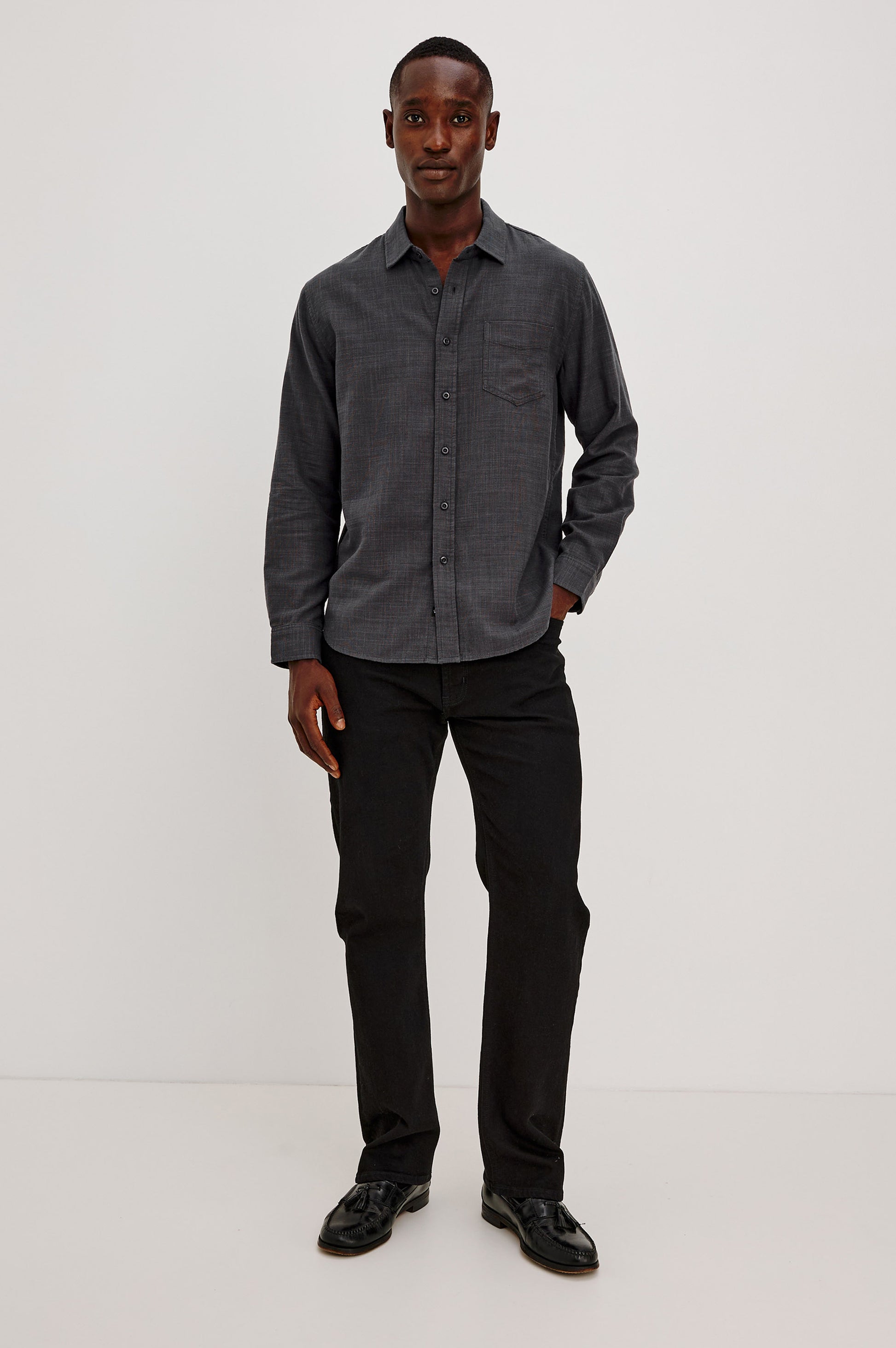 A man stands against a plain white background, wearing the Rails Wyatt Long Sleeve Button Down shirt in dark gray cotton with a relaxed fit, paired with black pants and black shoes. He has one hand in his pocket and smiles slightly at the camera.