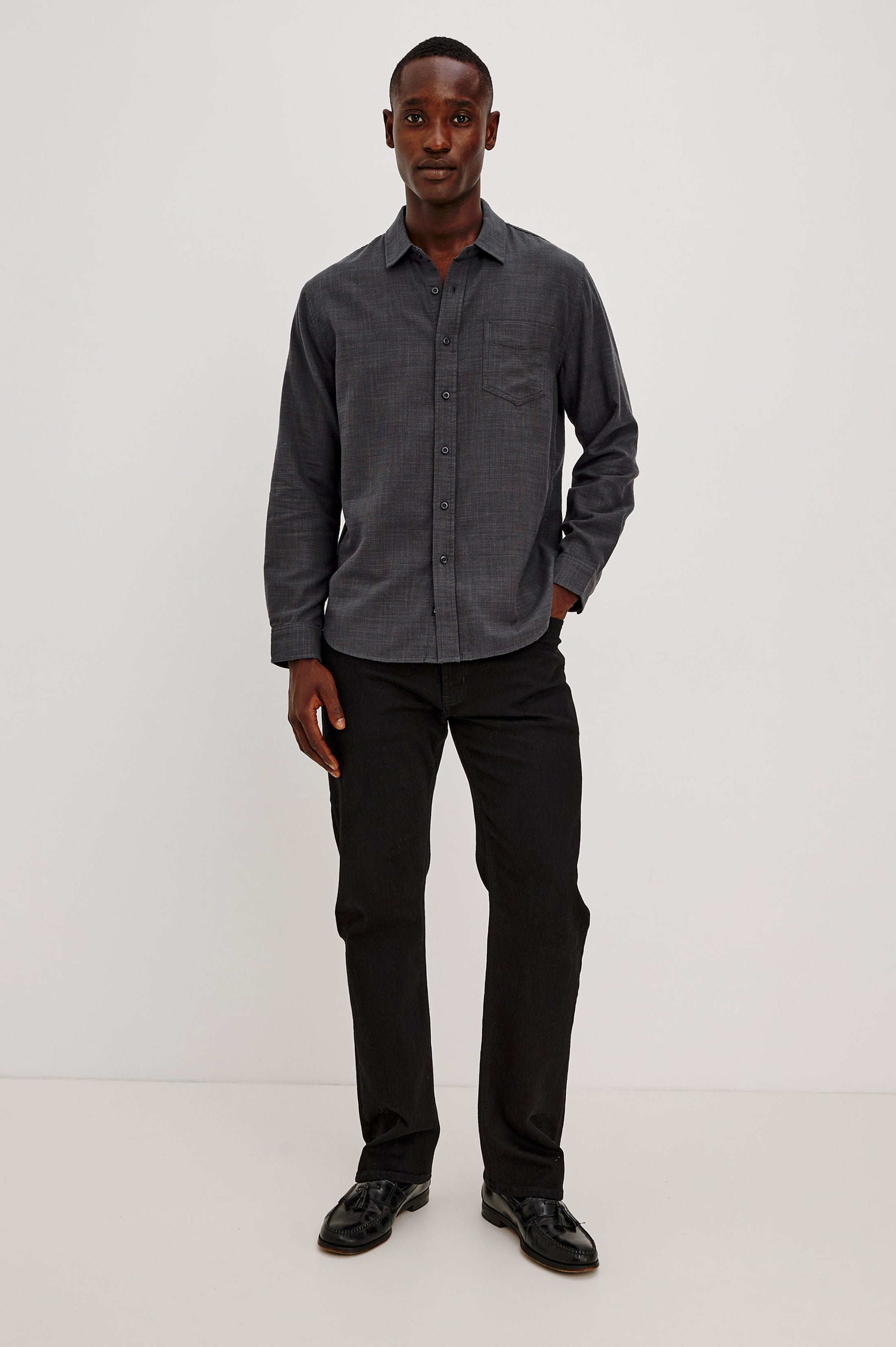 A man stands against a plain white background, wearing the Rails Wyatt Long Sleeve Button Down shirt in dark gray cotton with a relaxed fit, paired with black pants and black shoes. He has one hand in his pocket and smiles slightly at the camera.