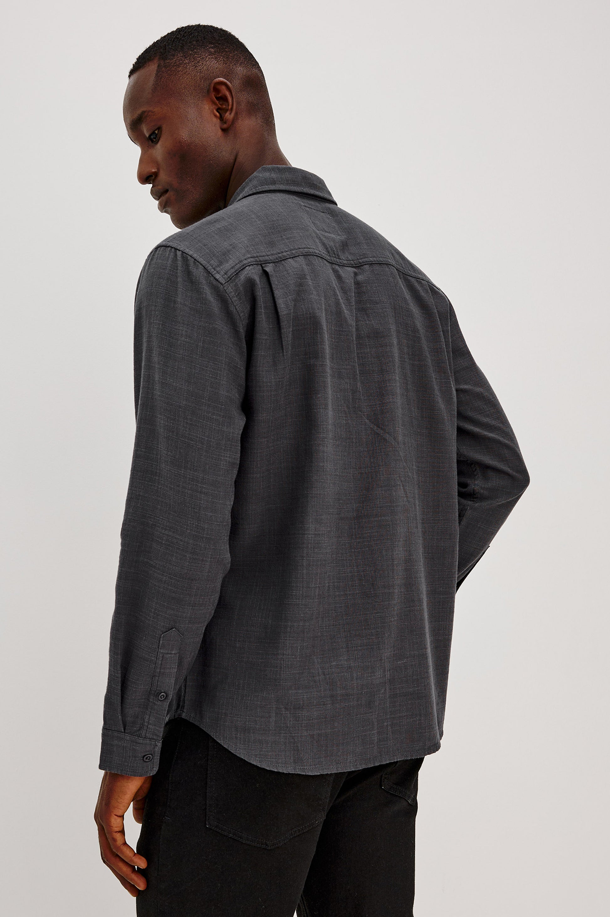 A man stands facing away, looking over his shoulder, wearing the Rails Wyatt Long Sleeve Button Down in dark grey cotton, paired with black pants against a plain light background.