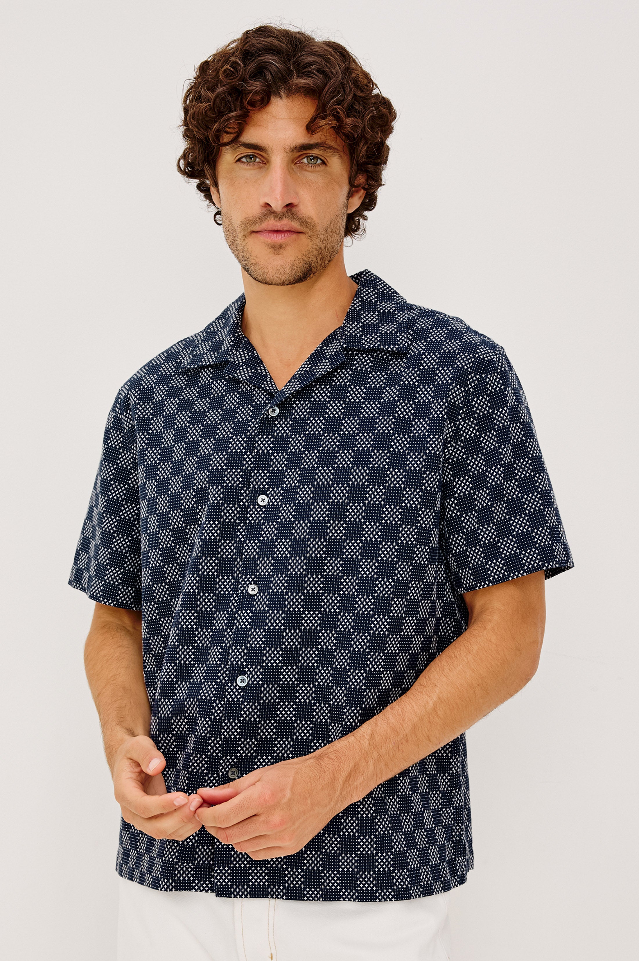 A man with curly brown hair and light facial hair wears the Rails Kin shirt in Navy Multi, featuring a relaxed fit and white geometric pattern, standing against a plain light background.