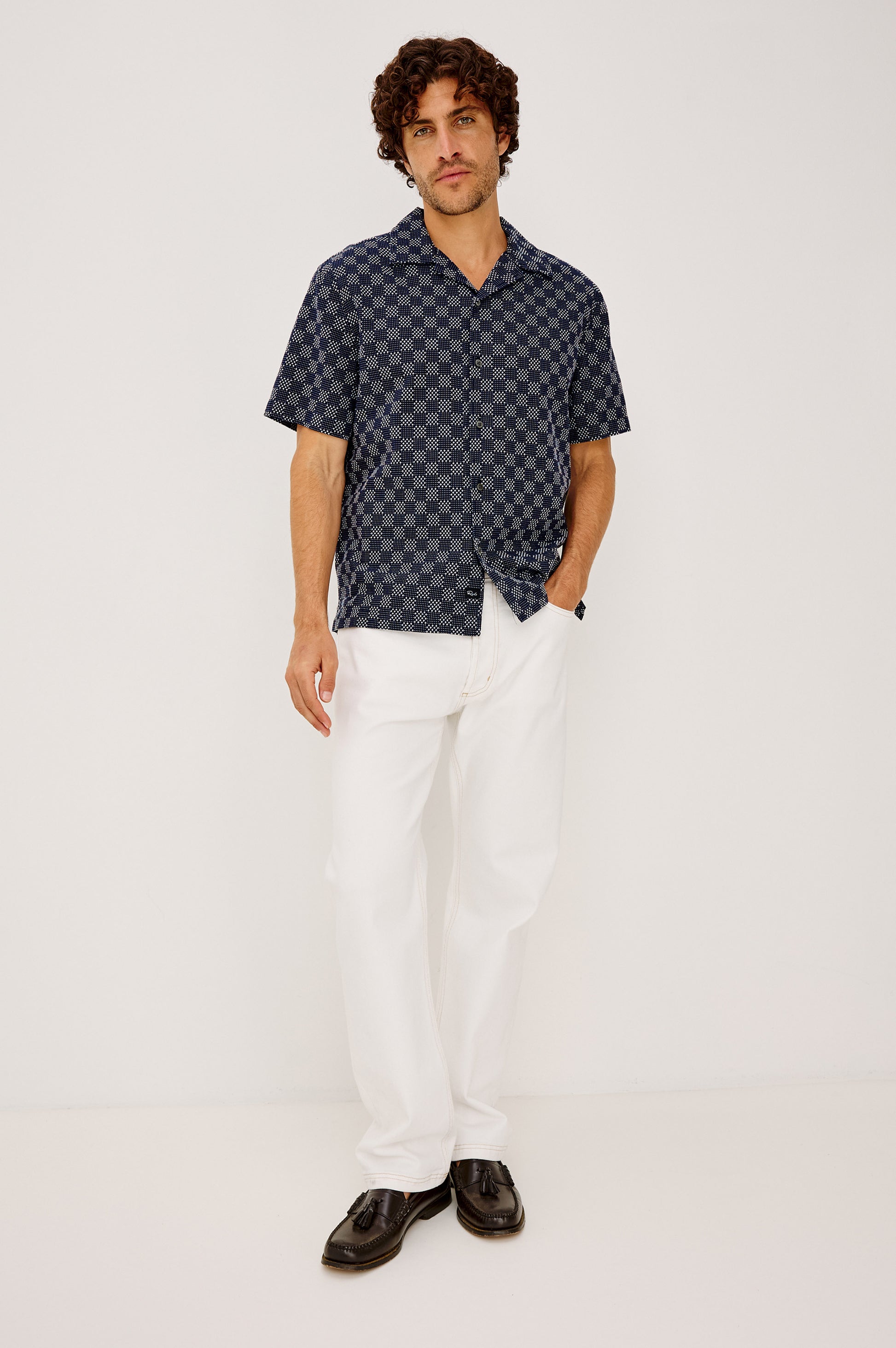 A man with curly brown hair and a neutral expression wears the Rails Kin relaxed fit, short-sleeve dark blue checkered camp collar shirt in textured dobby cotton, paired with white pants and black shoes against a plain white background.