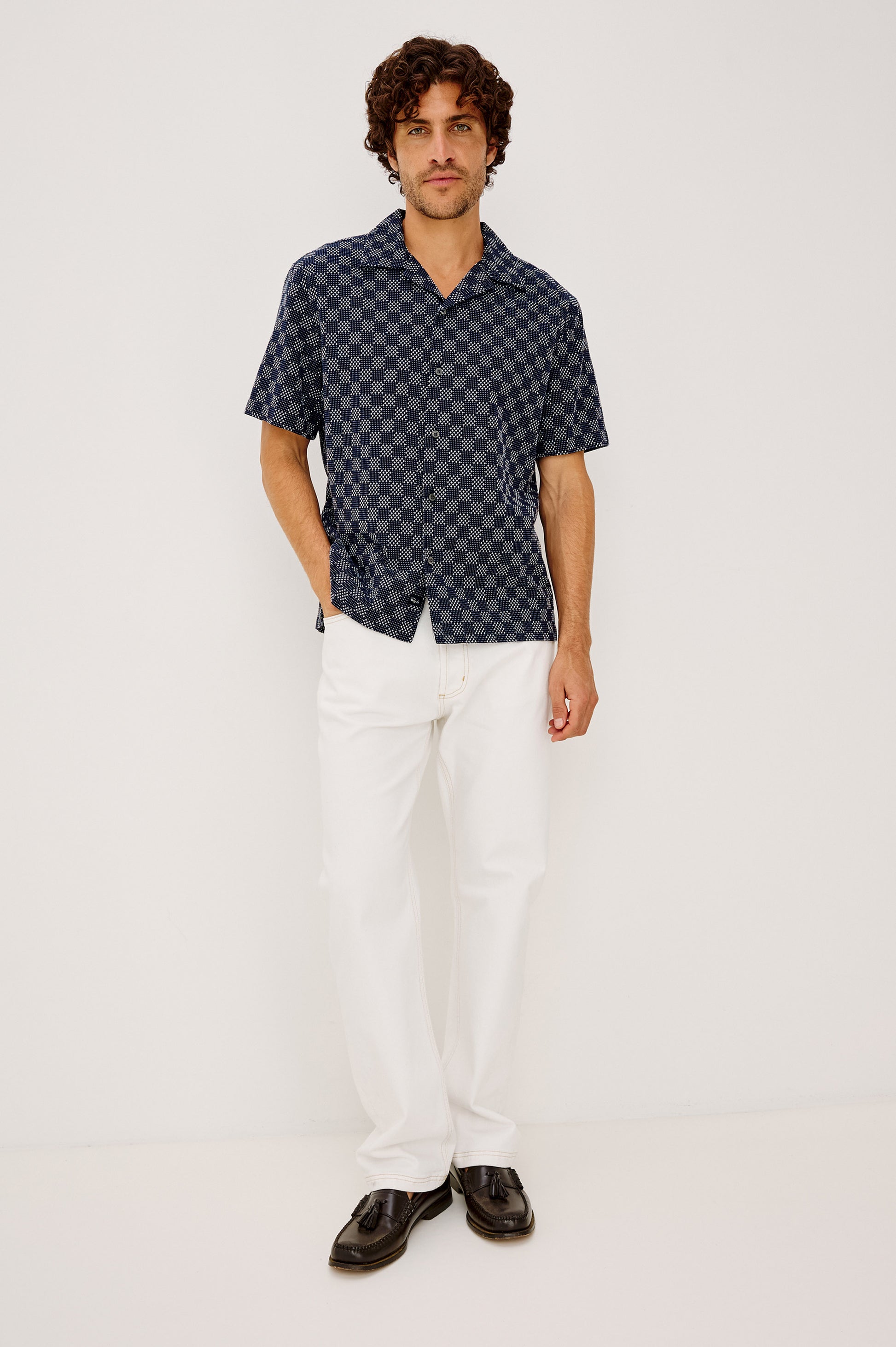 A man stands against a plain white background, wearing the Rails Kin relaxed fit navy checkered camp collar shirt, paired with white pants and black dress shoes, with one hand in his pocket.