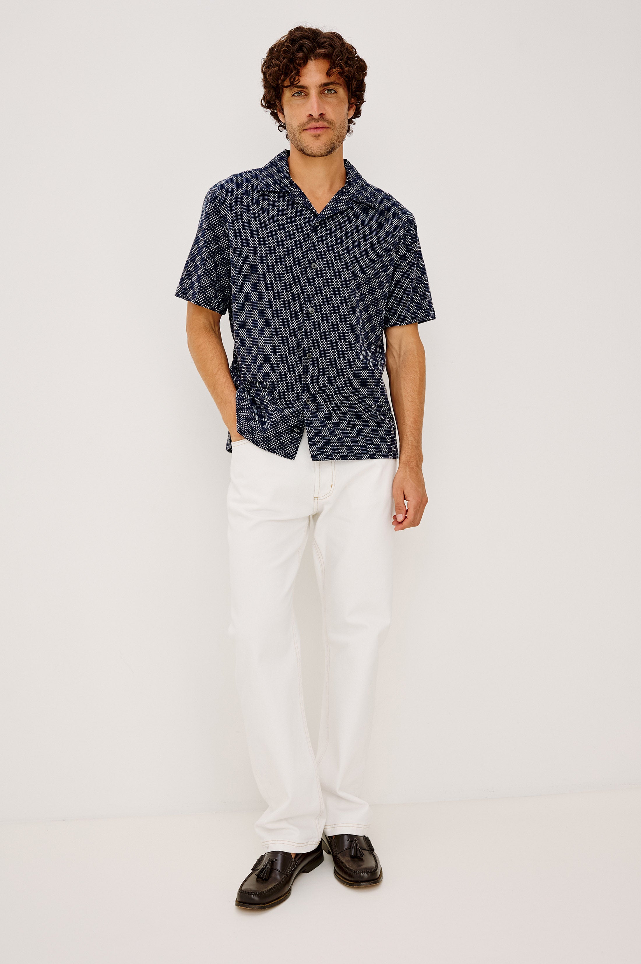 A man stands against a plain white background, wearing the Rails Kin relaxed fit navy checkered camp collar shirt, paired with white pants and black dress shoes, with one hand in his pocket.