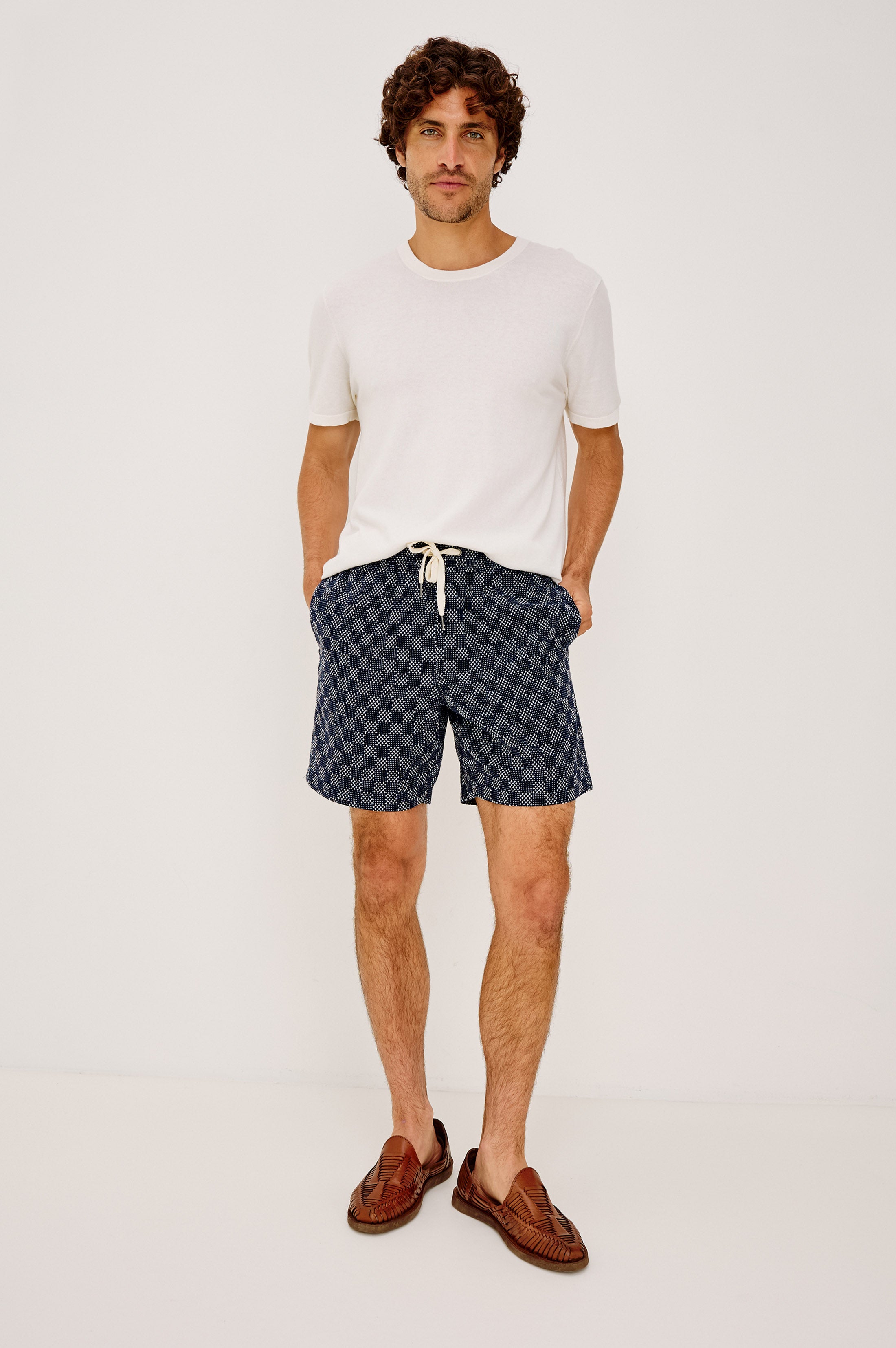 A man with curly hair stands against a white background wearing the Rails Cruz white short-sleeve t-shirt, blue relaxed-fit drawstring shorts, and brown woven loafers, with his hands in his pockets.