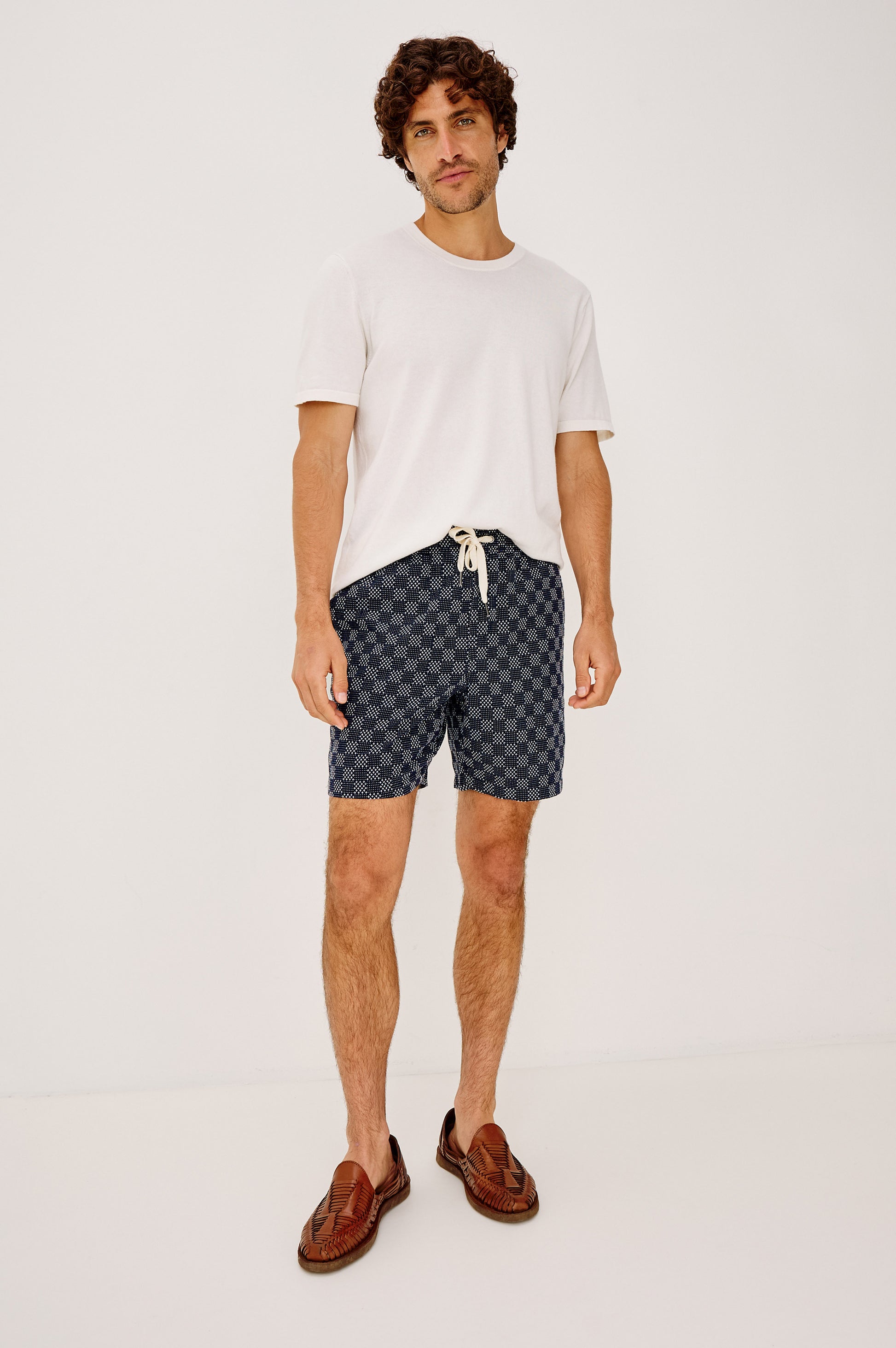 A man with curly hair and a relaxed expression wears the Rails Cruz white t-shirt, relaxed fit blue check shorts with a drawstring, and brown woven loafers, standing against a plain white background.