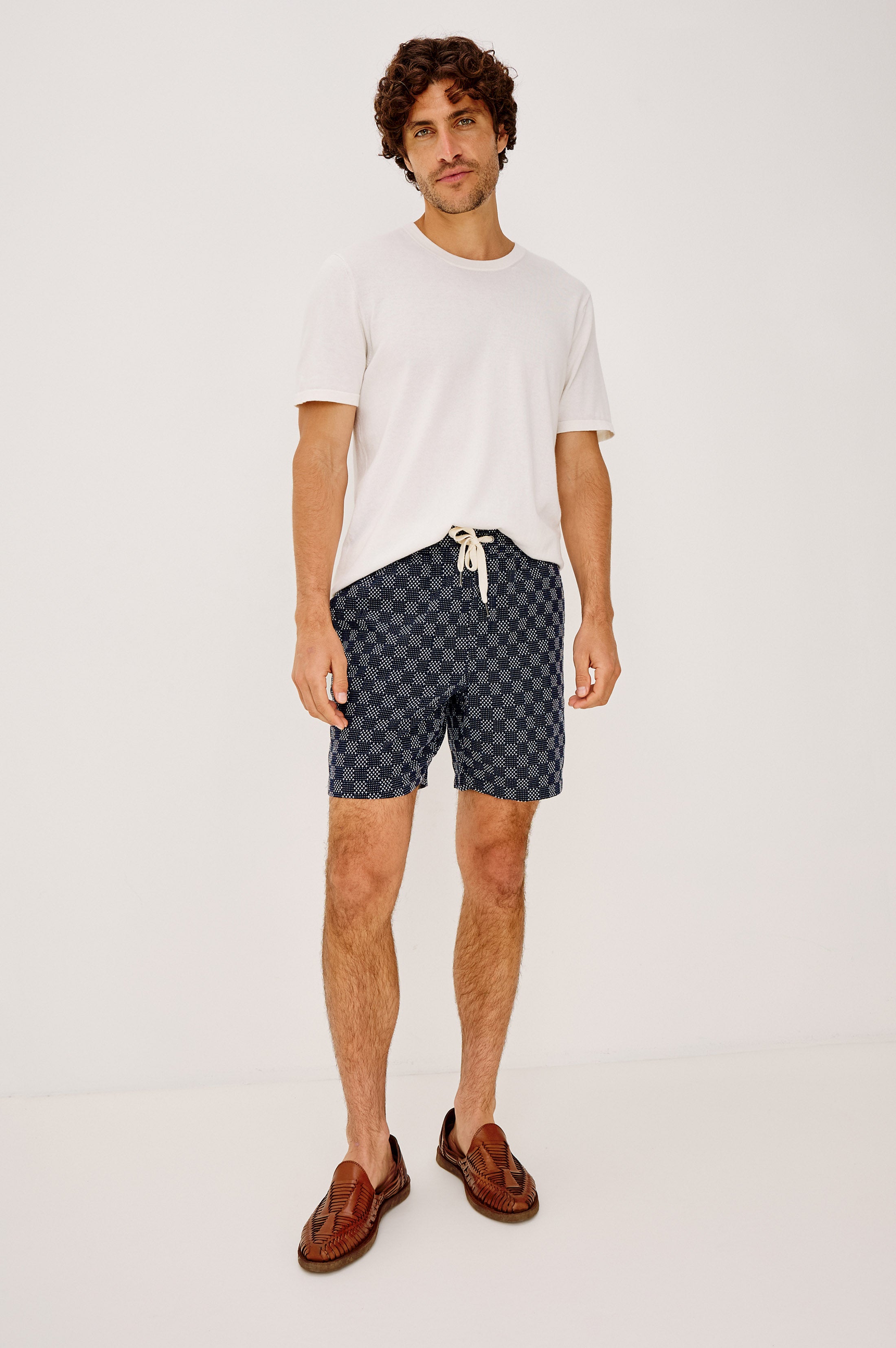 A man with curly hair and a relaxed expression wears the Rails Cruz white t-shirt, relaxed fit blue check shorts with a drawstring, and brown woven loafers, standing against a plain white background.