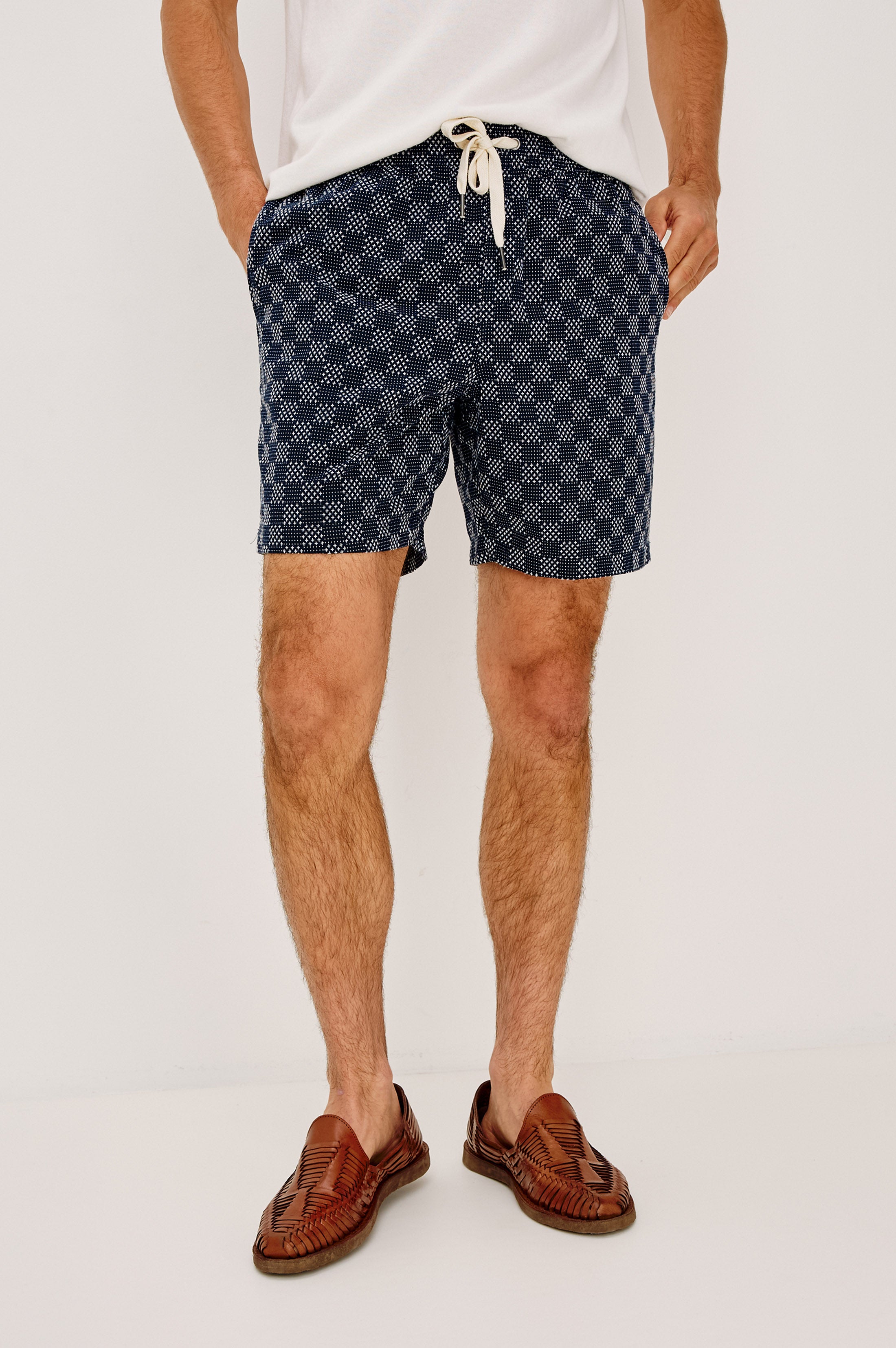 A man stands against a plain background, wearing the Rails Cruz relaxed fit shorts in Navy Multi with a white drawstring, paired with a white shirt and brown loafers. His hands are in his pockets, and his face is out of frame.