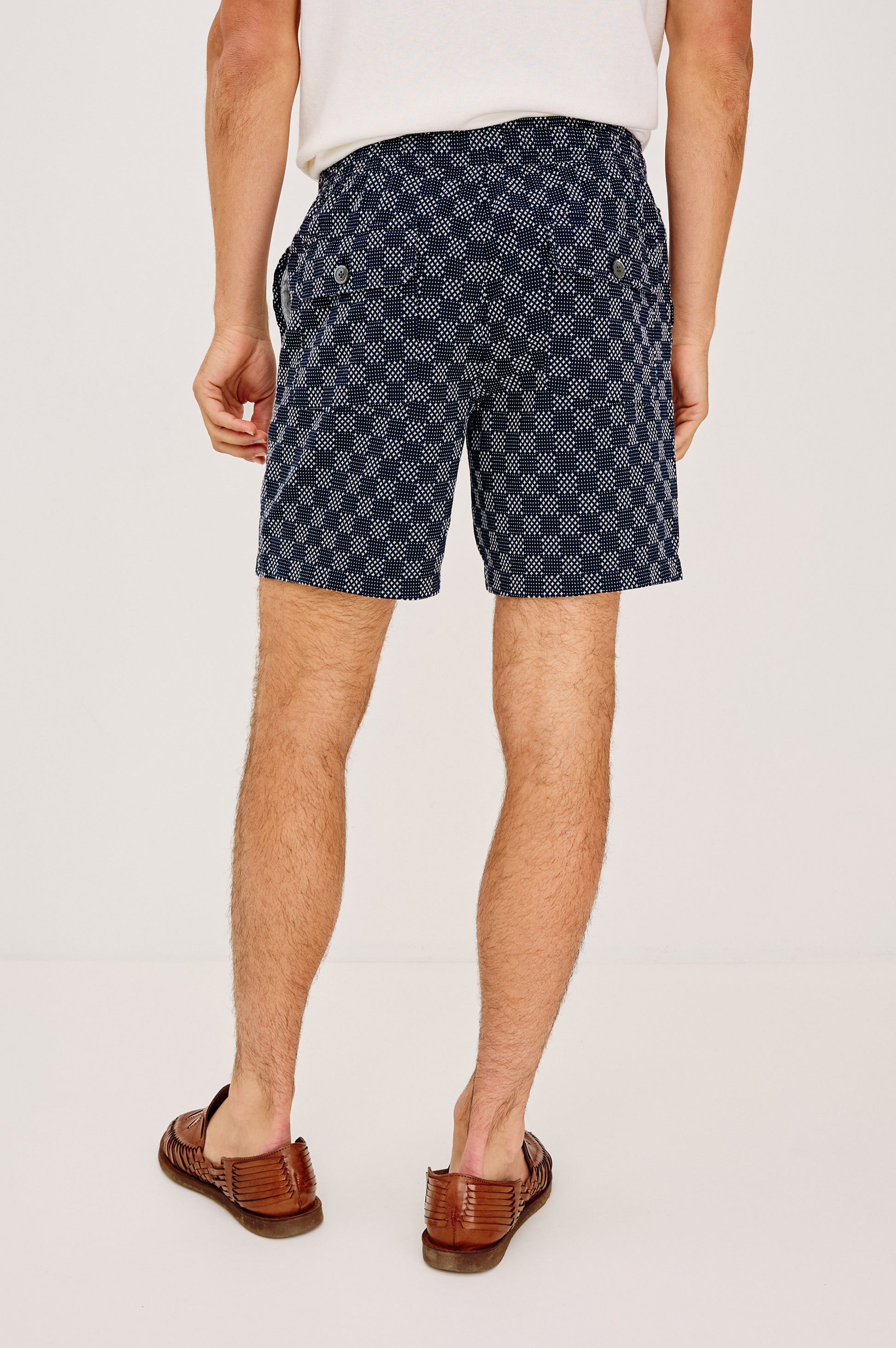 A man stands facing away in a white shirt, navy blue Cruz relaxed fit shorts with back pocket buttons by Rails, and brown woven loafers, against a plain white background.