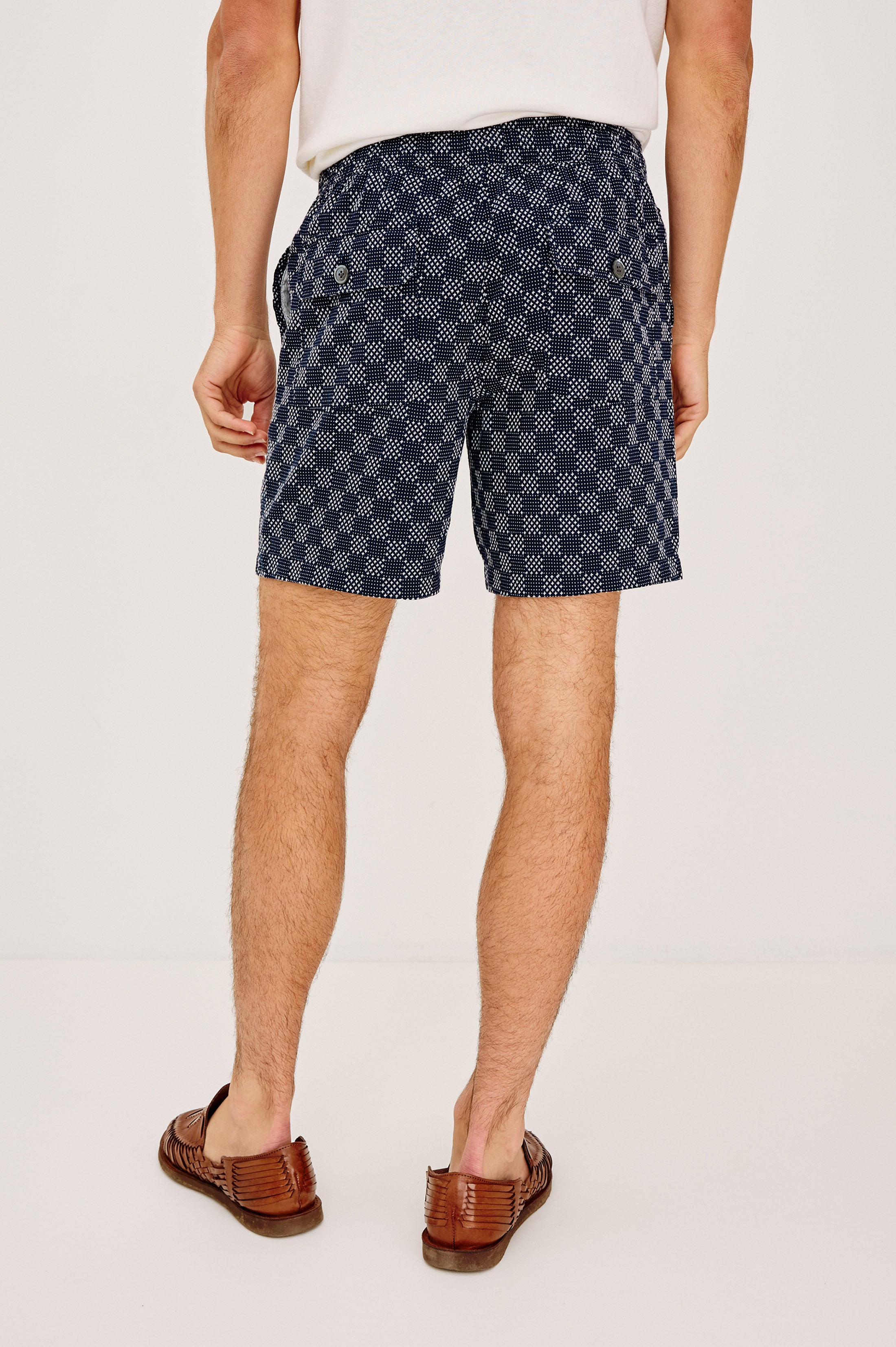 A man stands facing away in a white shirt, navy blue Cruz relaxed fit shorts with back pocket buttons by Rails, and brown woven loafers, against a plain white background.