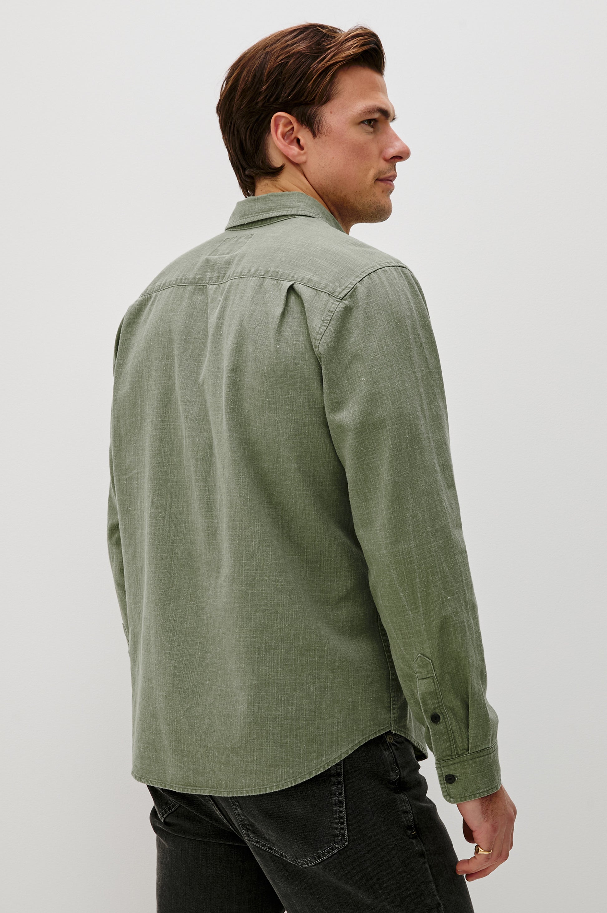 A man with brown hair wears the Rails Tristan Button Down Shirt in olive green, a relaxed fit, 100% cotton long-sleeve, paired with black jeans. He stands facing away against a plain white background. Machine wash cold.
