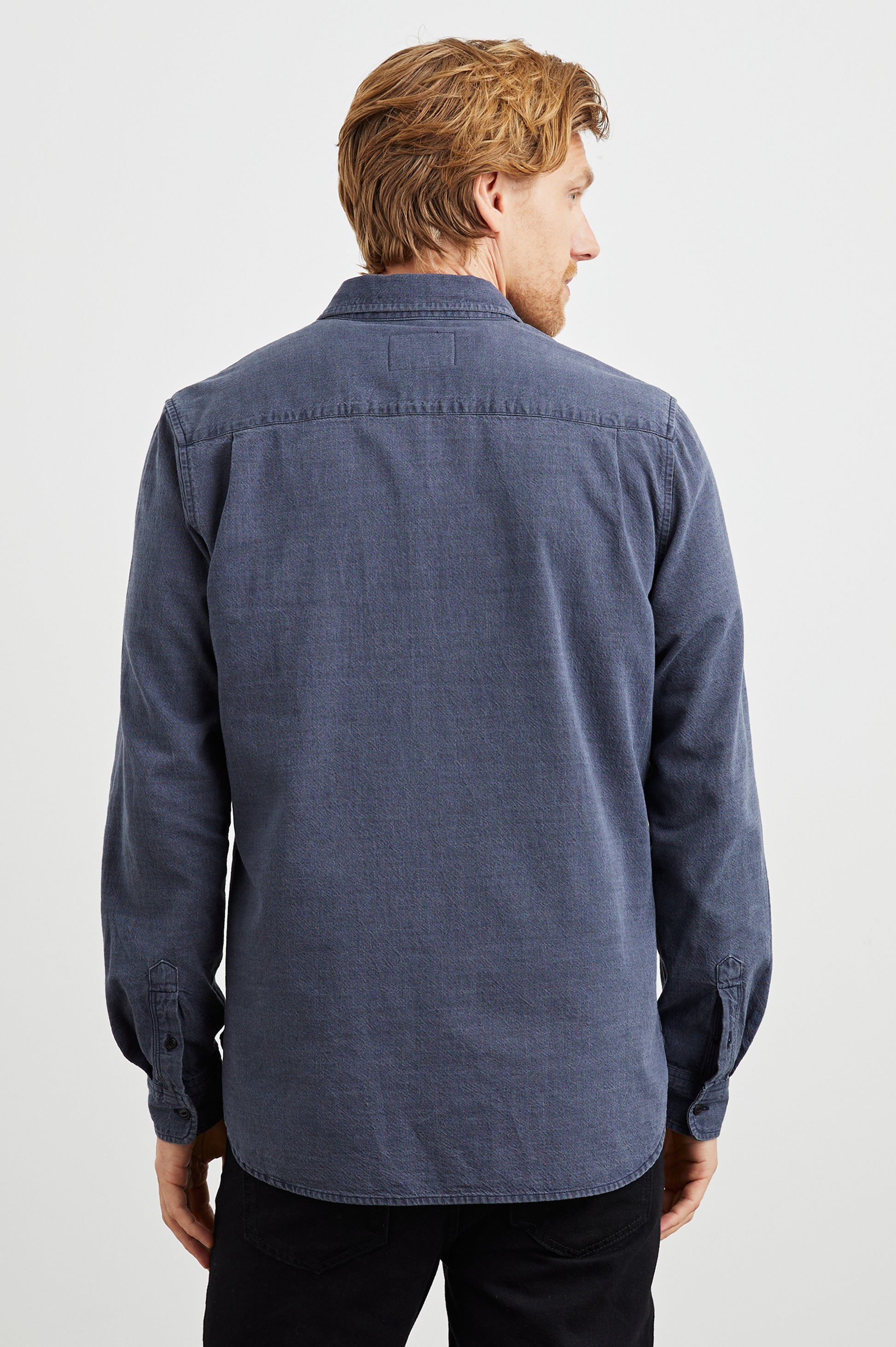 A man with light brown hair, seen from behind, wears the Rails Tristan Button Down Shirt—a relaxed fit, long-sleeved blue 100% cotton shirt—paired with black pants against a plain white background.