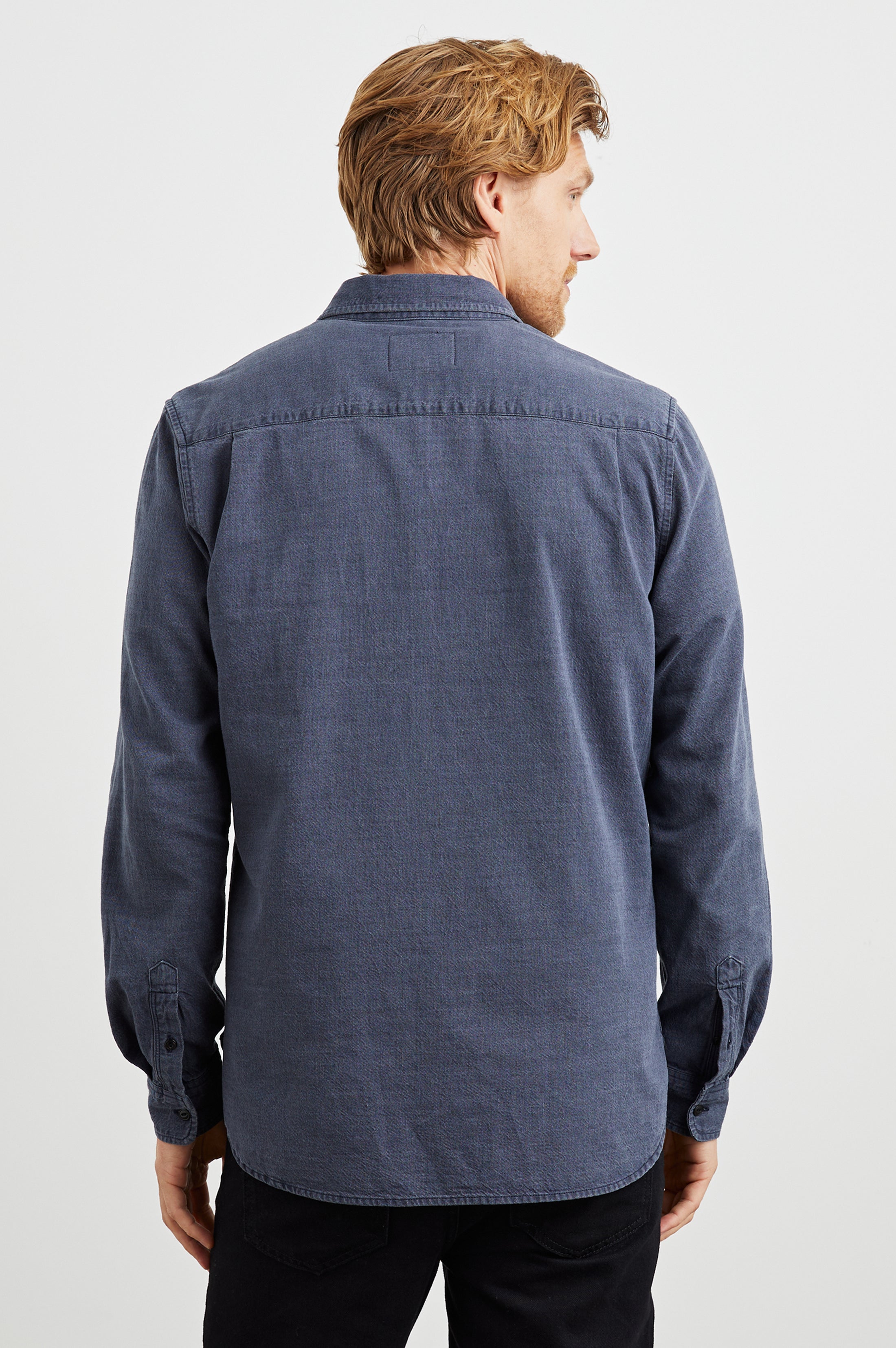A man with light brown hair, seen from behind, wears the Rails Tristan Button Down Shirt—a relaxed fit, long-sleeved blue 100% cotton shirt—paired with black pants against a plain white background.