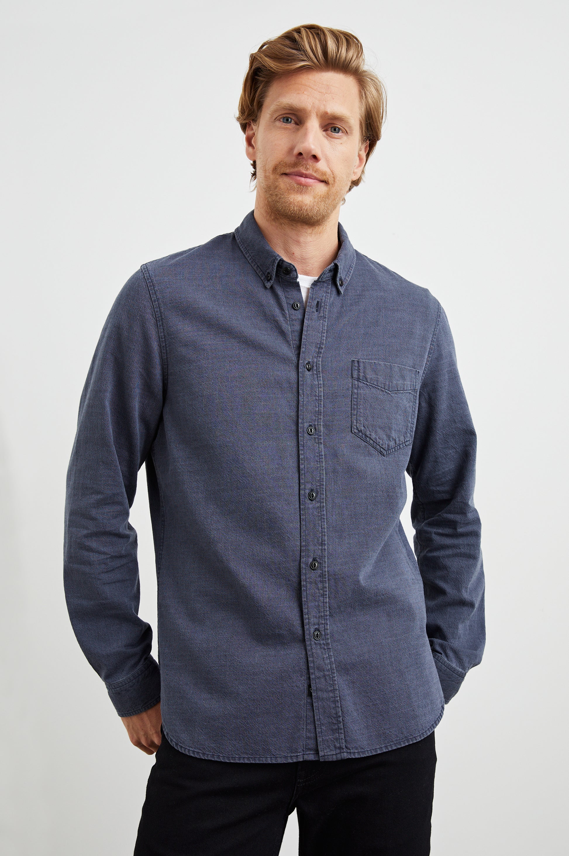A man with light brown hair and a short beard wears the Rails Tristan Button Down Shirt in blue, made from 100% cotton with a relaxed fit, paired with black pants. He stands confidently against a plain white background.