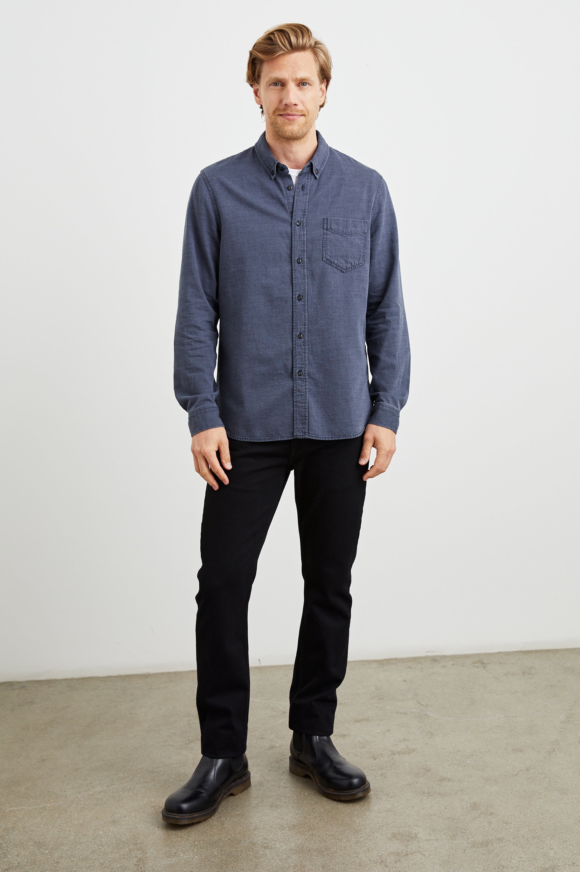 A man with short light brown hair stands indoors against a white wall, wearing the Rails Tristan Button Down Shirt in blue, paired with black pants and black boots. He faces the camera with a neutral expression.