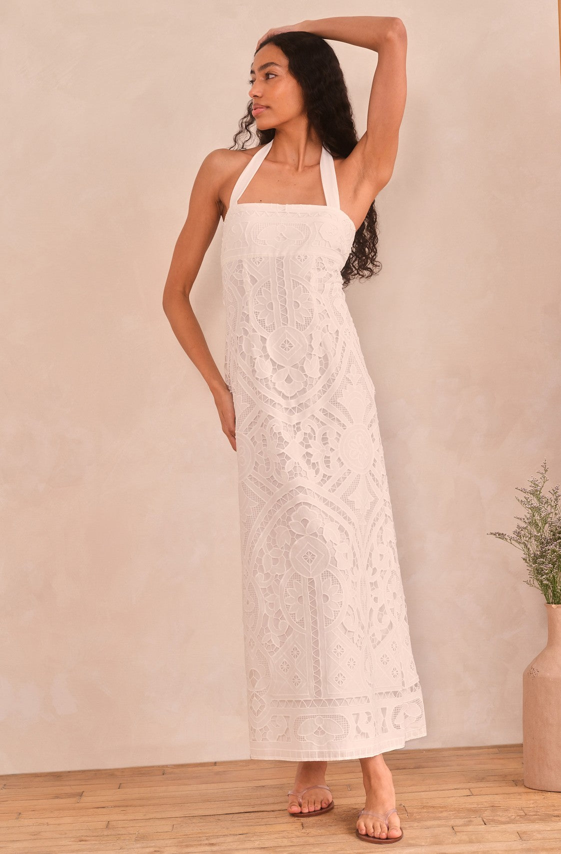 A woman poses barefoot indoors in the Saylor Rona Maxi Dress, a long white halter-neck style with intricate lace and an empire waist. She stands near a vase of greenery, one arm bent behind her head as she looks to the side.