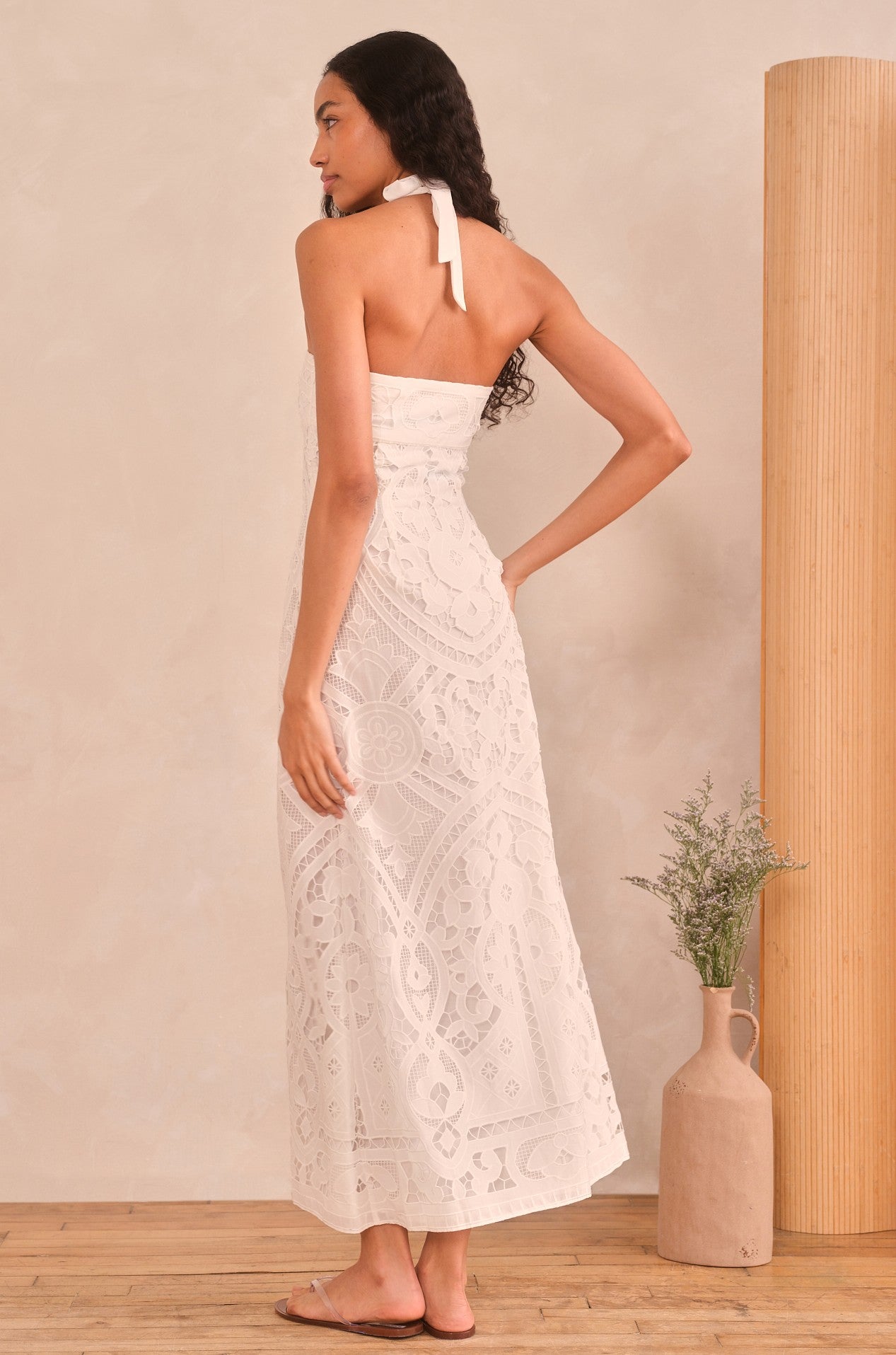 A woman with long, wavy hair wears the Saylor Rona Maxi Dress—a white lace 100% cotton gown—with sandals, standing indoors on a wooden floor near a tall vase of greenery and a cylindrical column. She faces away, glancing sideways.