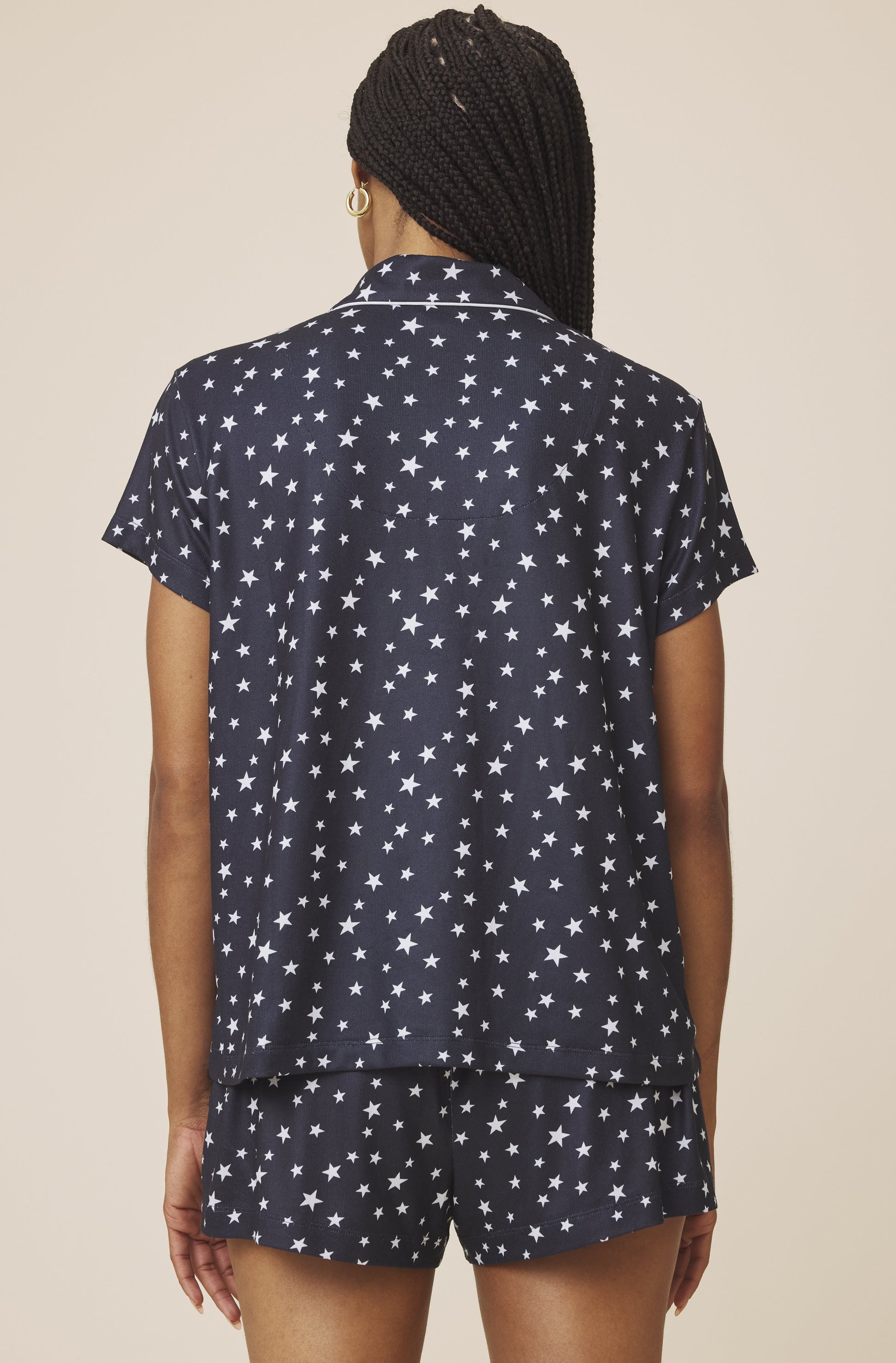 A person with braided hair is seen from behind wearing the Splendid Flora Pillow Soft PJ Short Set, a women's navy blue pajama set with white stars, featuring a short-sleeve button-up top and matching shorts against a beige background.