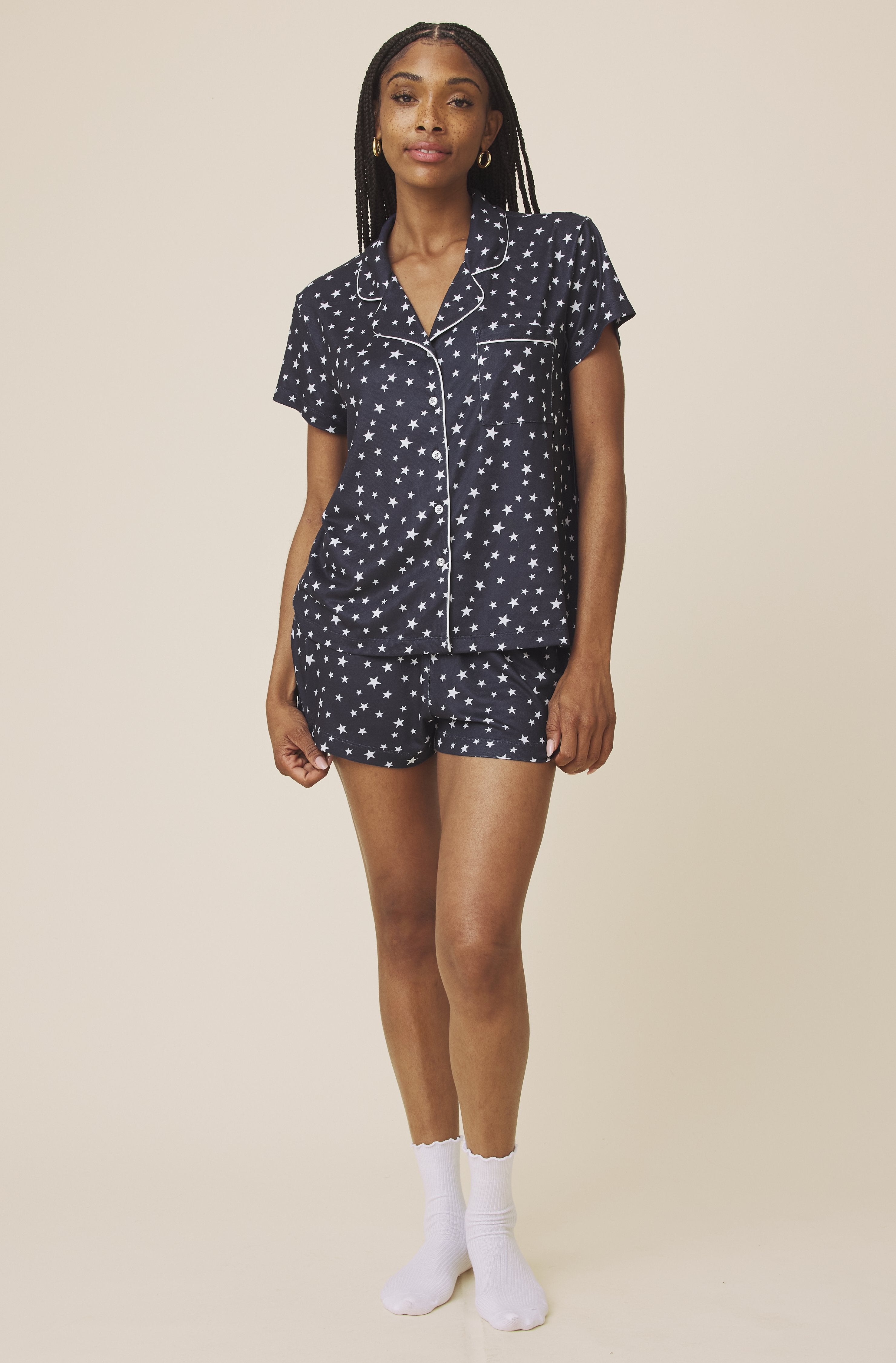 A woman with long braided hair stands against a plain background, wearing the Splendid Flora Pillow Soft PJ Short Set—navy blue with white star patterns and trim—while looking at the camera in this lightweight sleepwear.